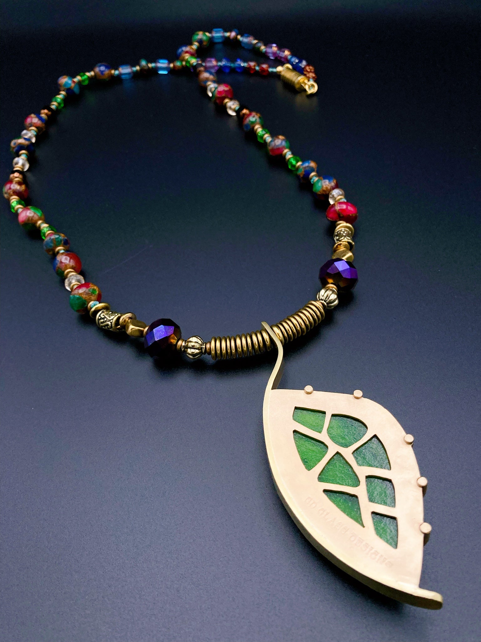 Back of dichroic glass set in a unique brass pendant showcasing the unique cutouts, revealing the green glass beneath. Jewelers brass setting has prongs on left side and a textured bezel on the right side. Pendant is hung on a 29" long hand-knotted multicolor beaded necklace, by 3D Glass Designs.