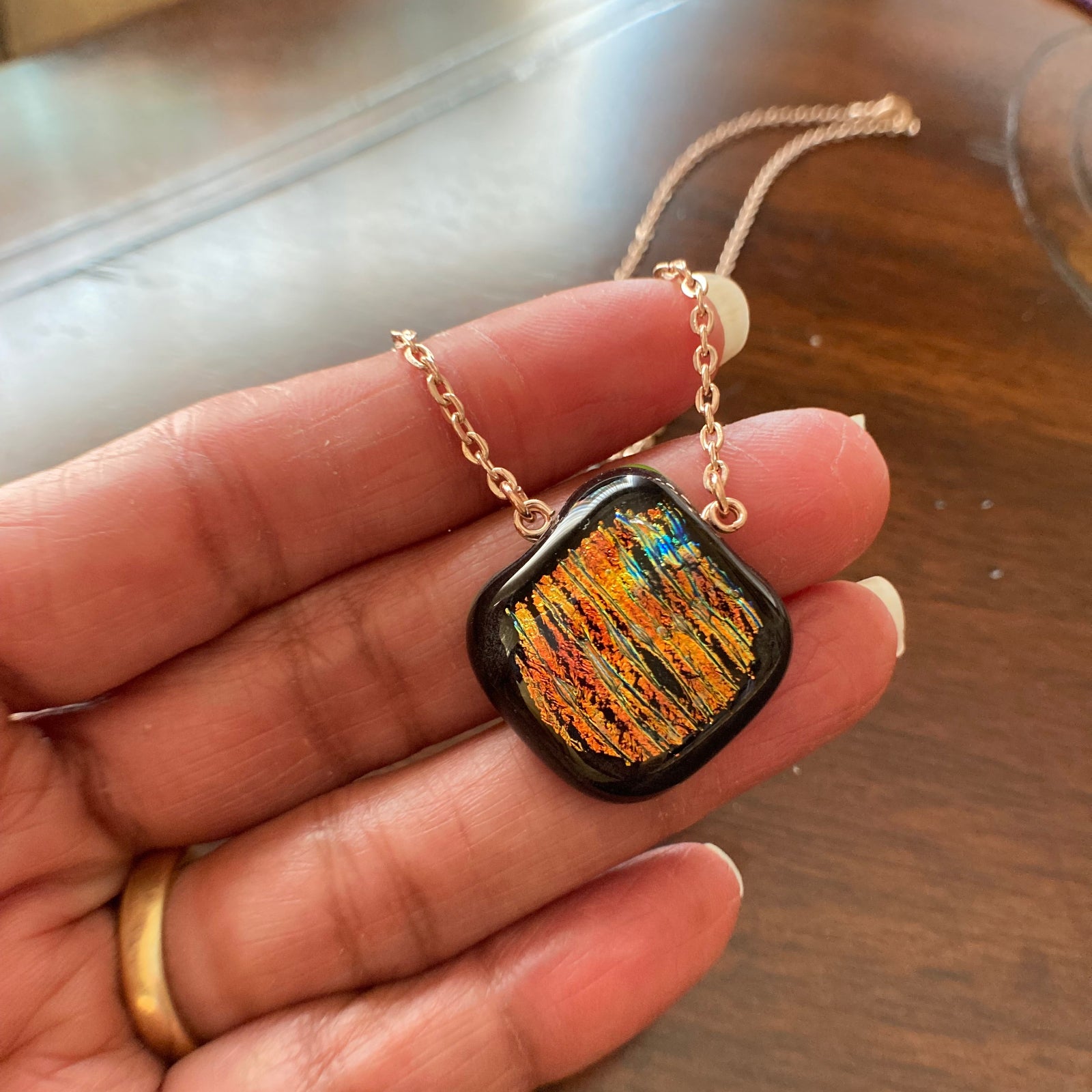 Hand holding orange fused glass pendant against a wooden table, by 3D Glass Designs.