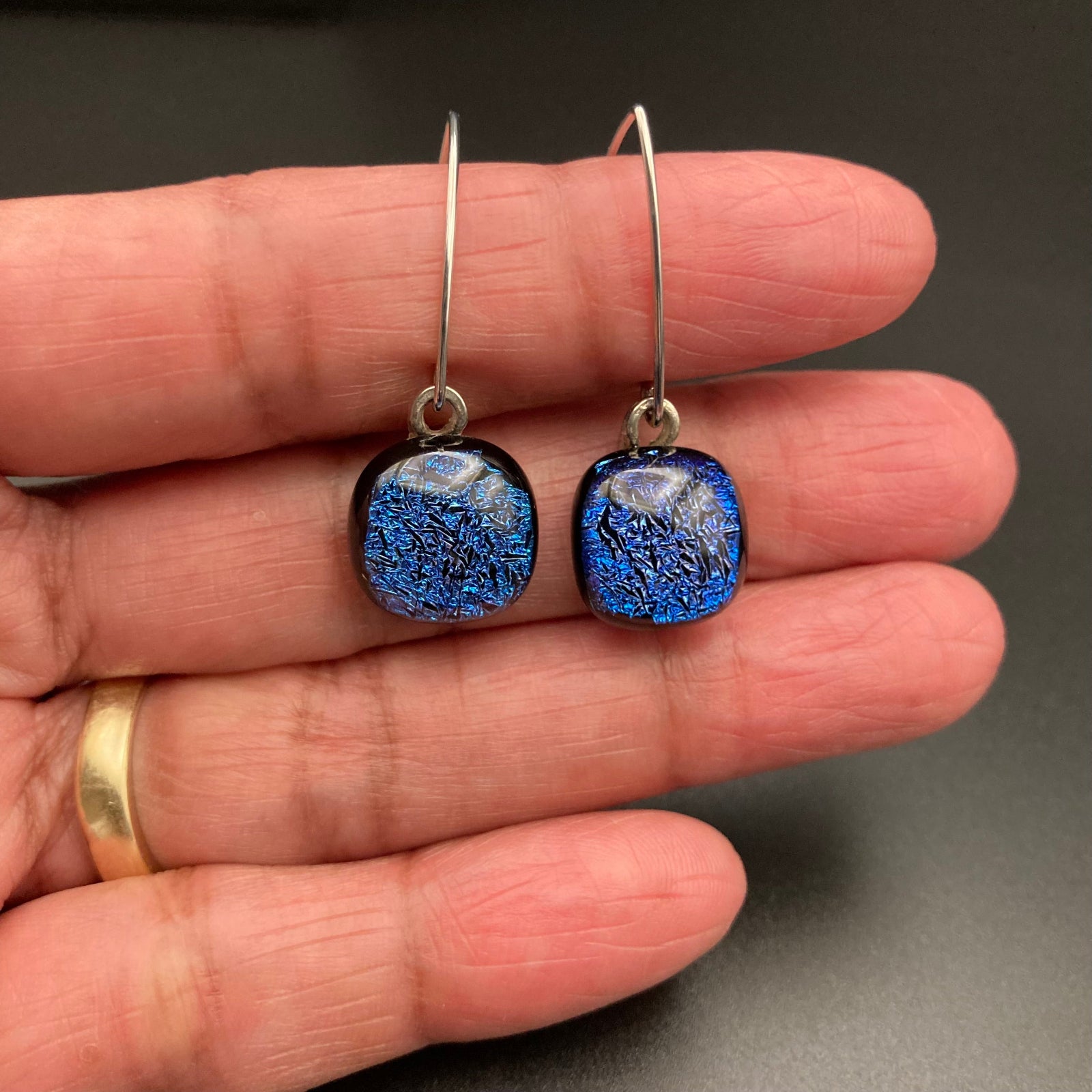 A hand holding a pair of earrings with blue dangles and silver ear wires, by 3D Glass Designs.