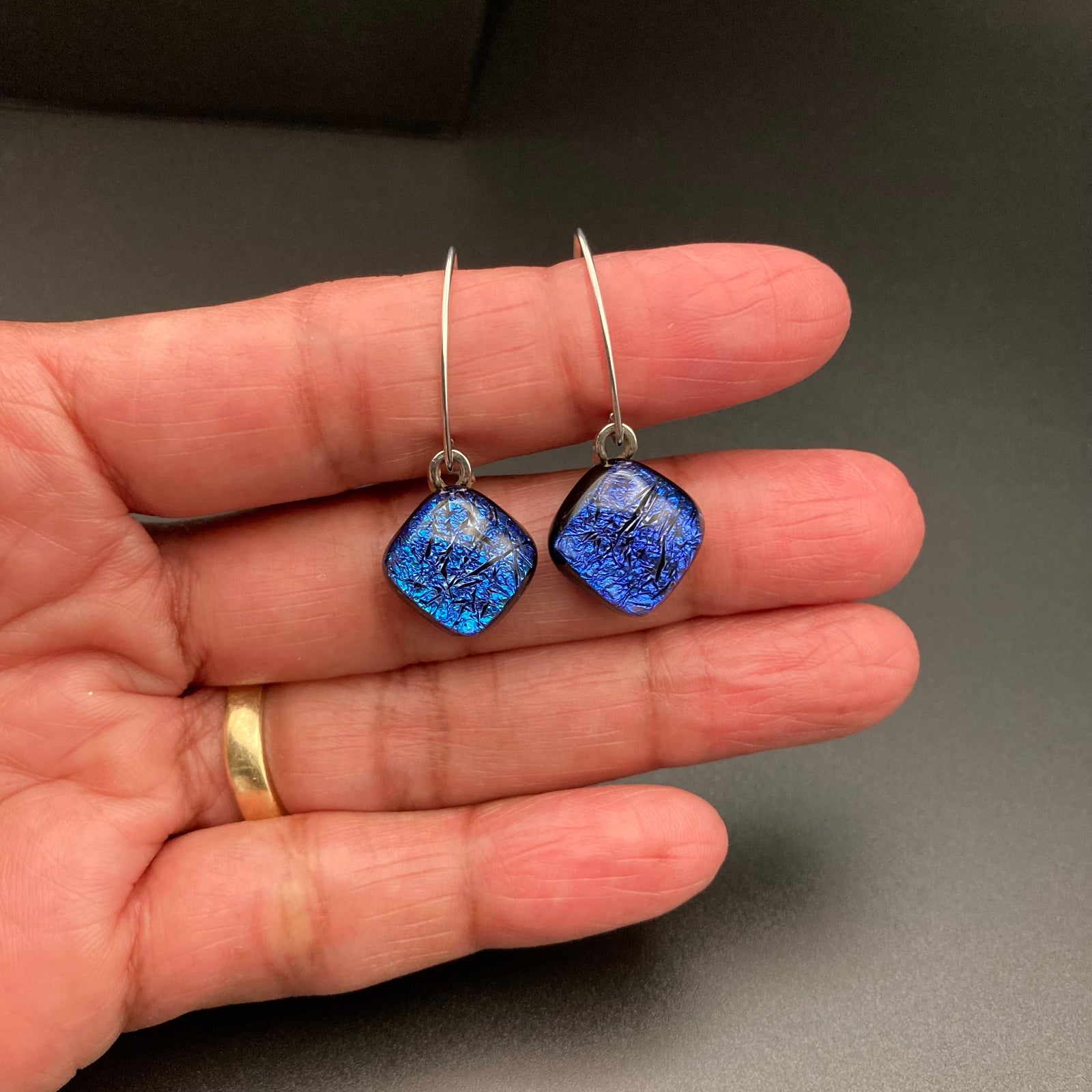 A hand holding a pair of fused glass earrings with diamond-shaped drops, by 3D Glass Designs.