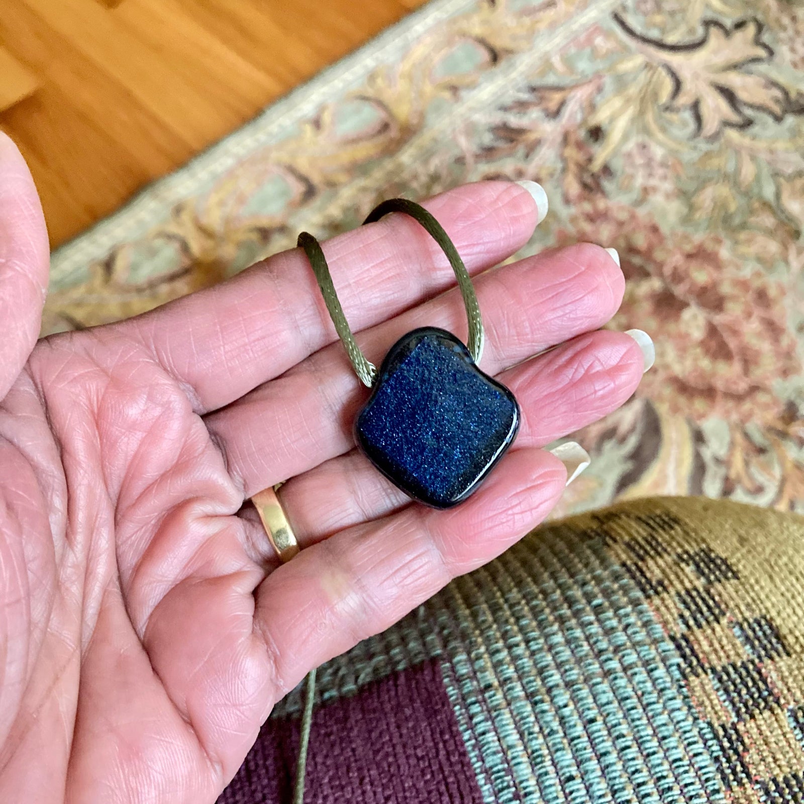 Hand holding green glass pendant on an olive-green satin cord against a sage floral rug, by 3D Glass Designs.