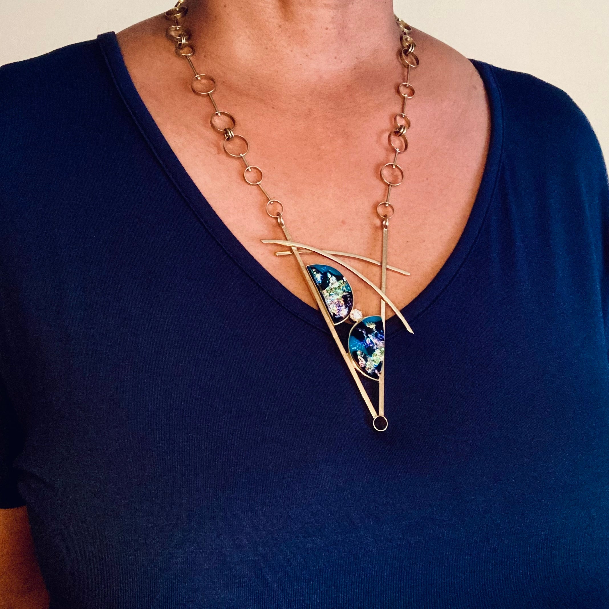 Model wearing geometric inspired brass pendant with teal fused glass in semi-circles and CZ, framed in a V with two slightly curved arch's above. Necklace made of various sized circles linked together, by 3D Glass Designs.