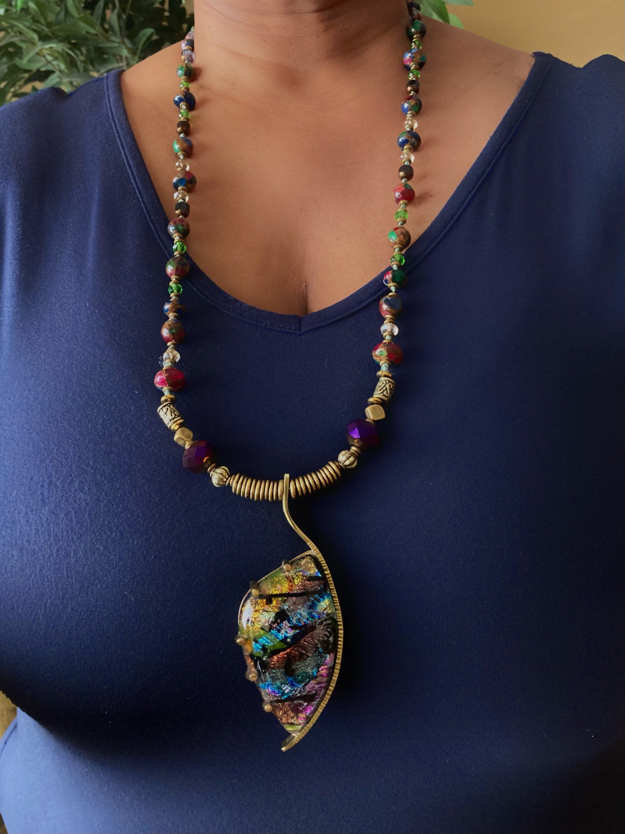 Model wearing dichroic glass set in a unique brass pendant, with prongs on left side and a textured bezel on the right side. Pendant is hung on a 29" long hand-knotted multicolor beaded necklace, by 3D Glass Designs.