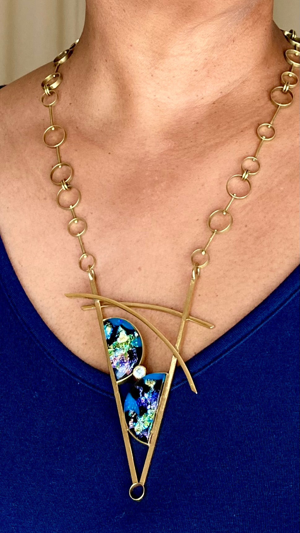 Model wearing brass necklace featuring two blue fused glass semi-circles, with a CZ in the middle, two slightly curved arch's above and a chain made of small, medium and large circles linked together, by 3D Glass Designs.