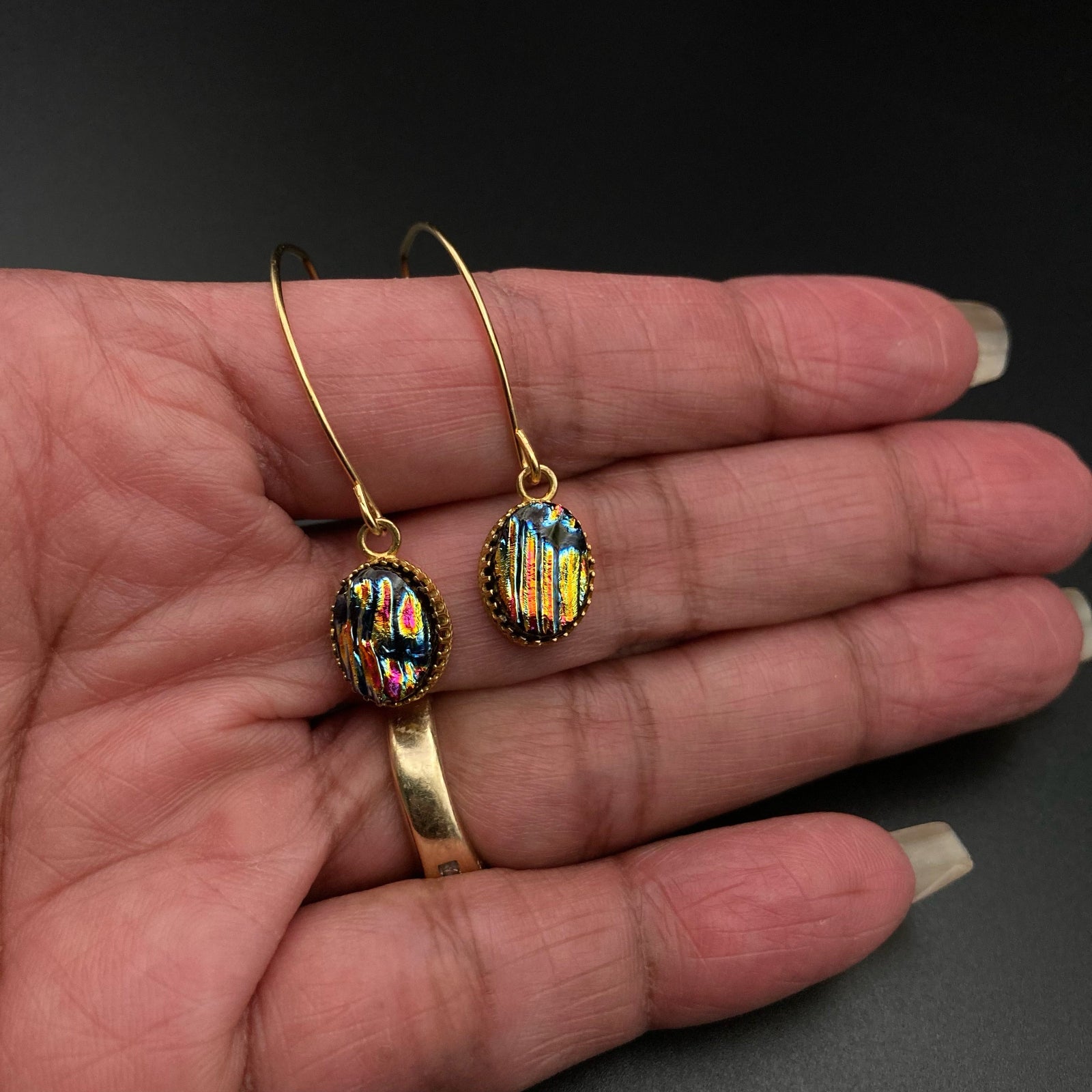 Hand holding a pair of pierced earrings with multi-colored dichroic glass drops on long gold-plated ear wires, by 3D Glass Designs.