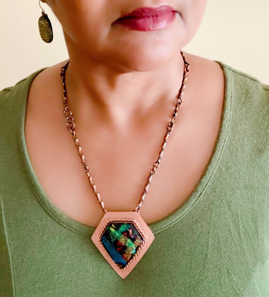 Model wearing diamond-shaped shield pendant necklace in fused glass and copper by 3D Glass Designs. Dichroic glass in blue, green and rust hues set in a handcrafted diamond-shaped shield copper pendant with an elegant rope detail surrounding the bezel. The handmade copper necklace consists of twisted copper links that resembles bamboo and a handmade clasp.