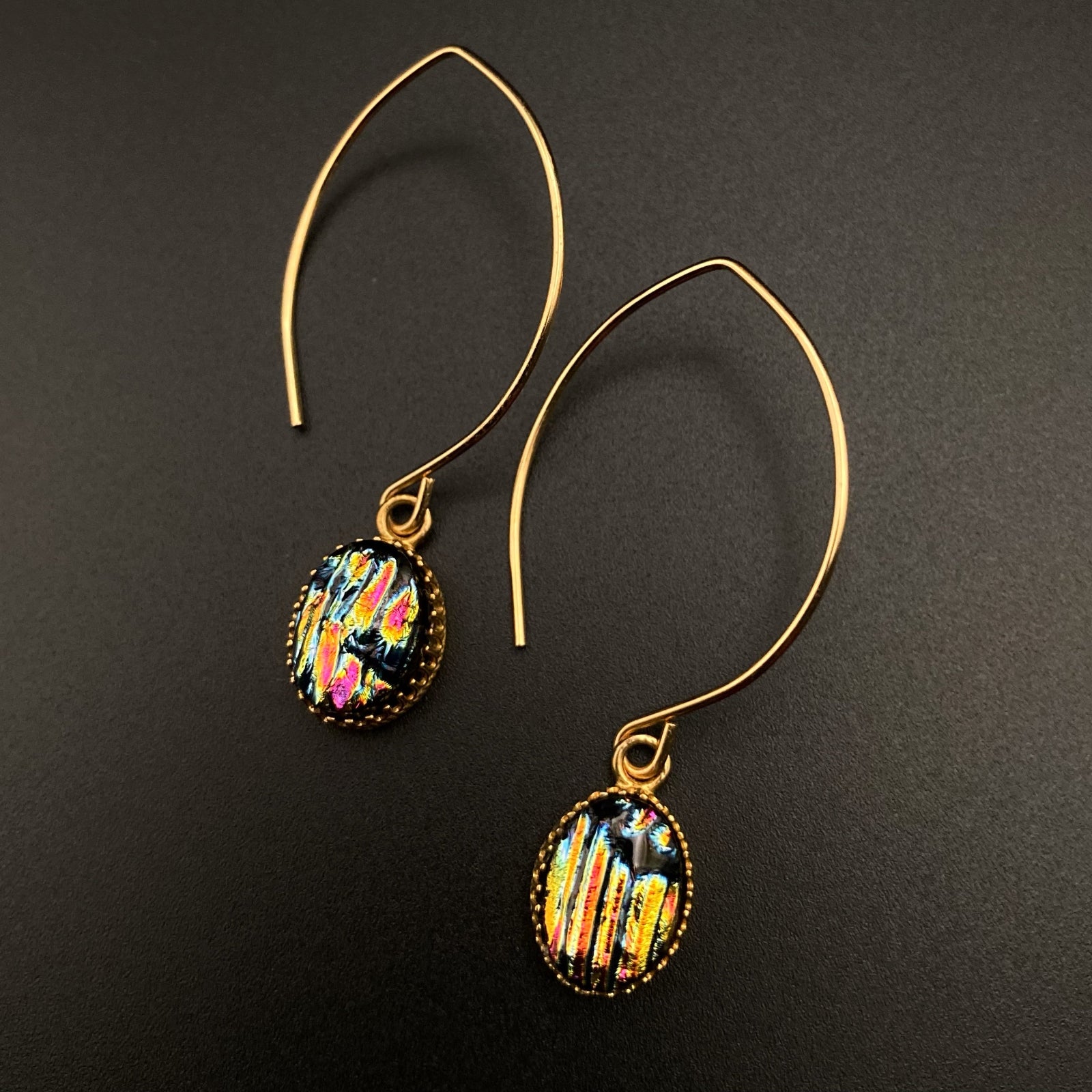 A pair of pierced earrings with multi-colored dichroic glass drops on long gold-plated ear wires against a grey background, by 3D Glass Designs.
