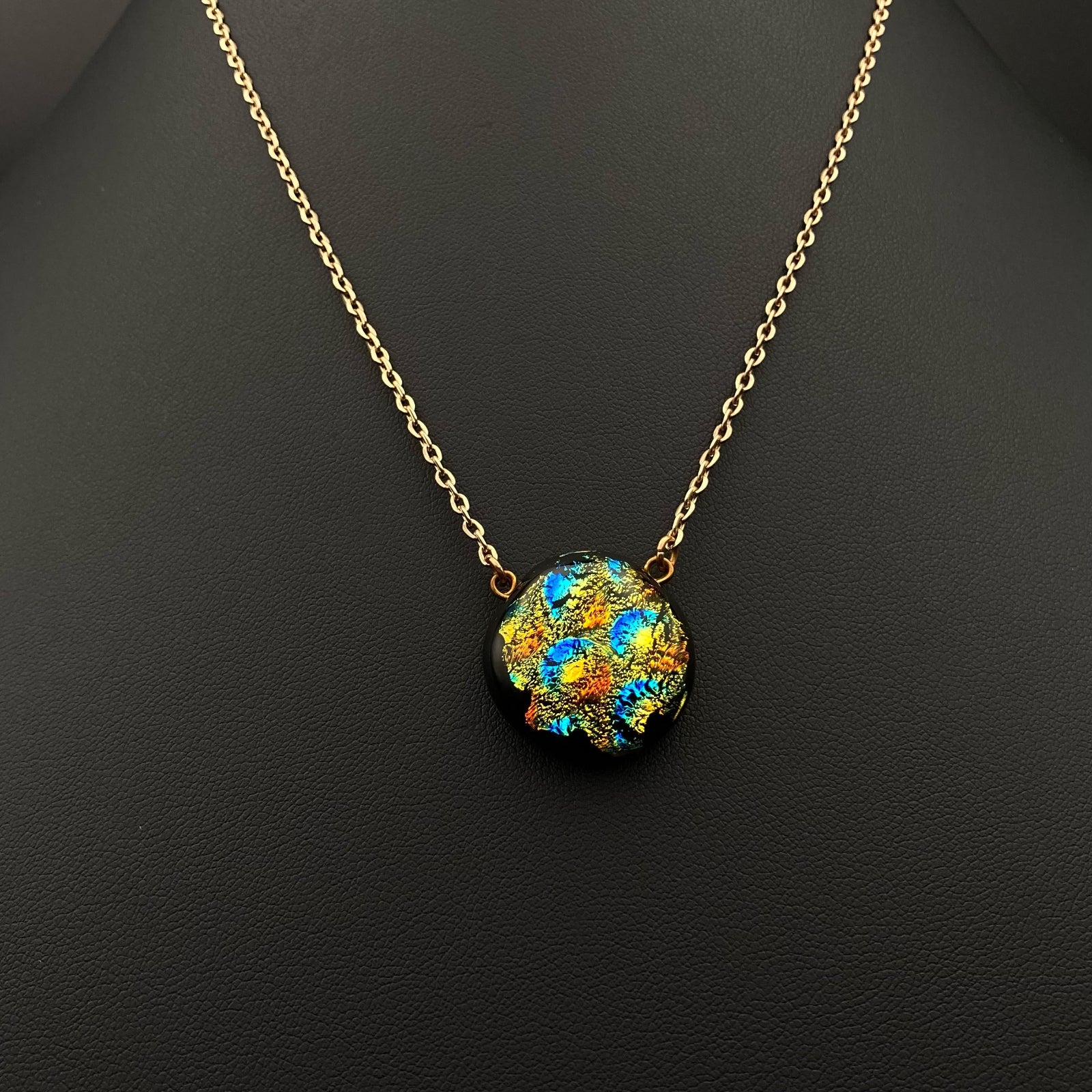 Colorful round fused glass pendant on a gold chain against a black leather background, by 3D Glass Designs.