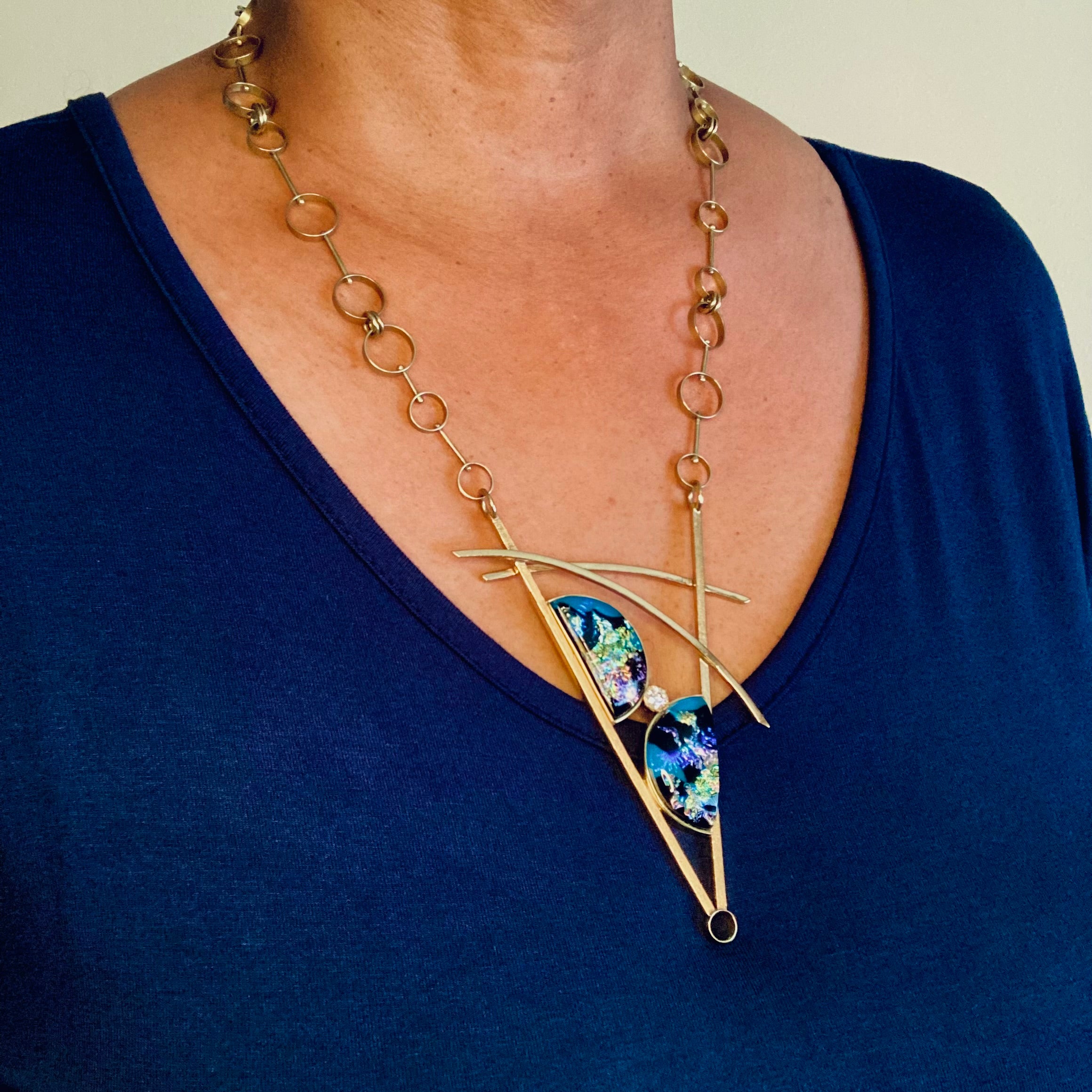 Model wearing geometric inspired brass pendant with teal fused glass in semi-circles and CZ, framed in a V with two slightly curved arch's above. Necklace made of various sized circles linked together, by 3D Glass Designs.
