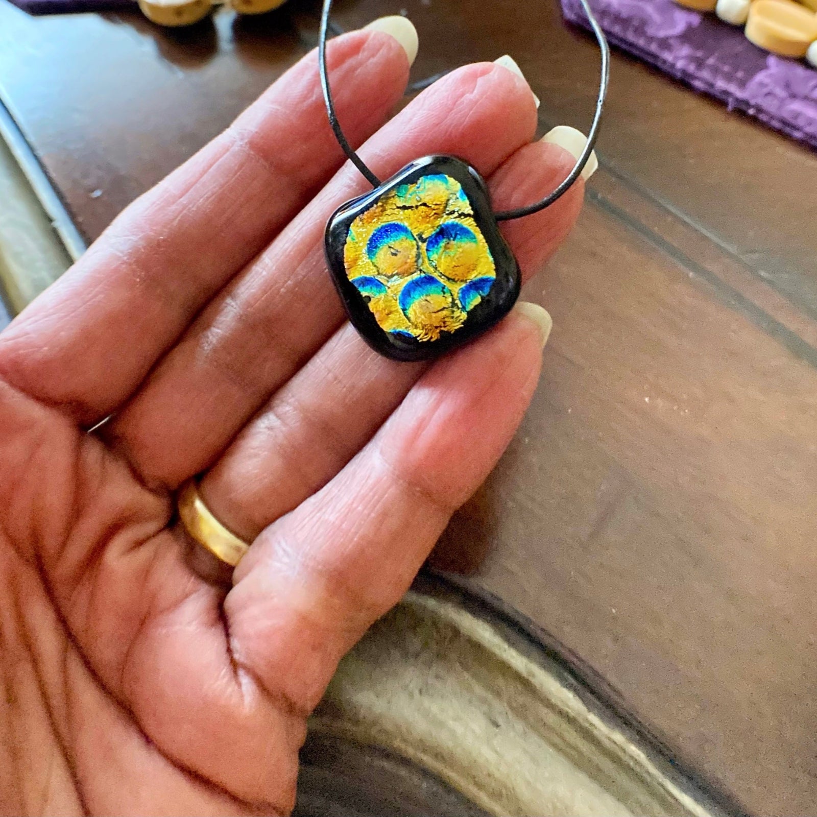 Hand holding dichroic glass pendant against a wooden tabletop, by 3D Glass Designs.