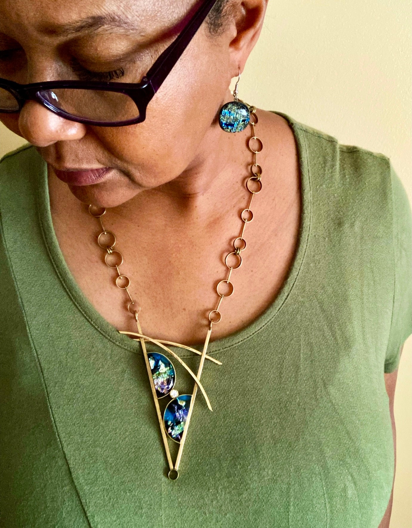 Model wearing geometric inspired brass pendant with teal fused glass in semi-circles and CZ, framed in a V with two slightly curved arch's above. Necklace made of various sized circles linked together, by 3D Glass Designs.