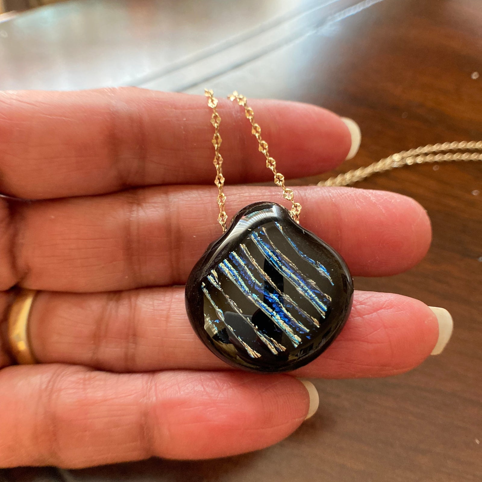 Hand holding dichroic glass pendant necklace against a wooden table, by 3D Glass Designs.