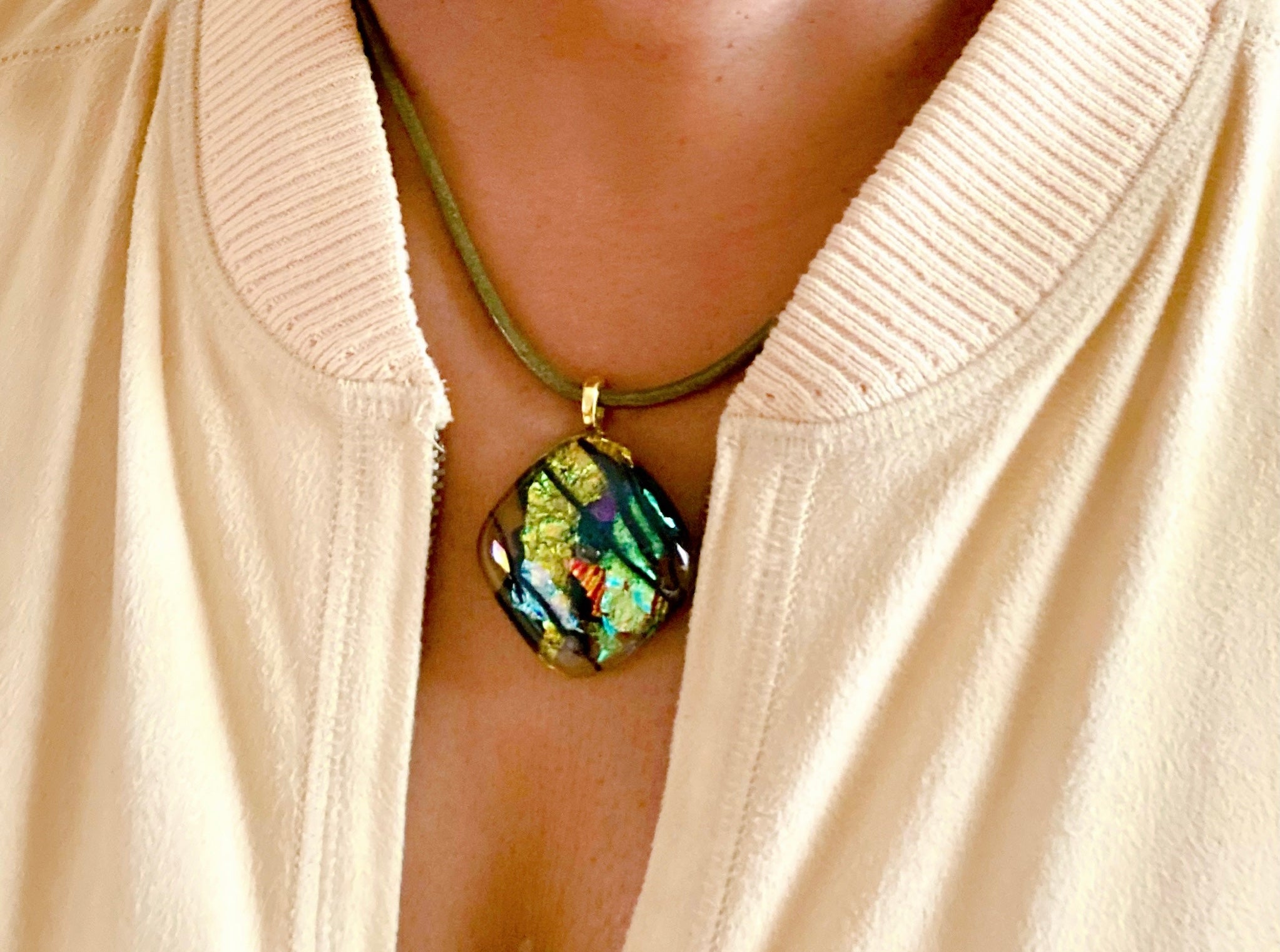 Model wearing fused glass pendant in transparent yellow with red, blue, green, purple and gold dichroic glass. Streaks of black add contrast to the design. Diamond shaped pendant hangs on an olive-green leather cord, by 3D Glass Designs.