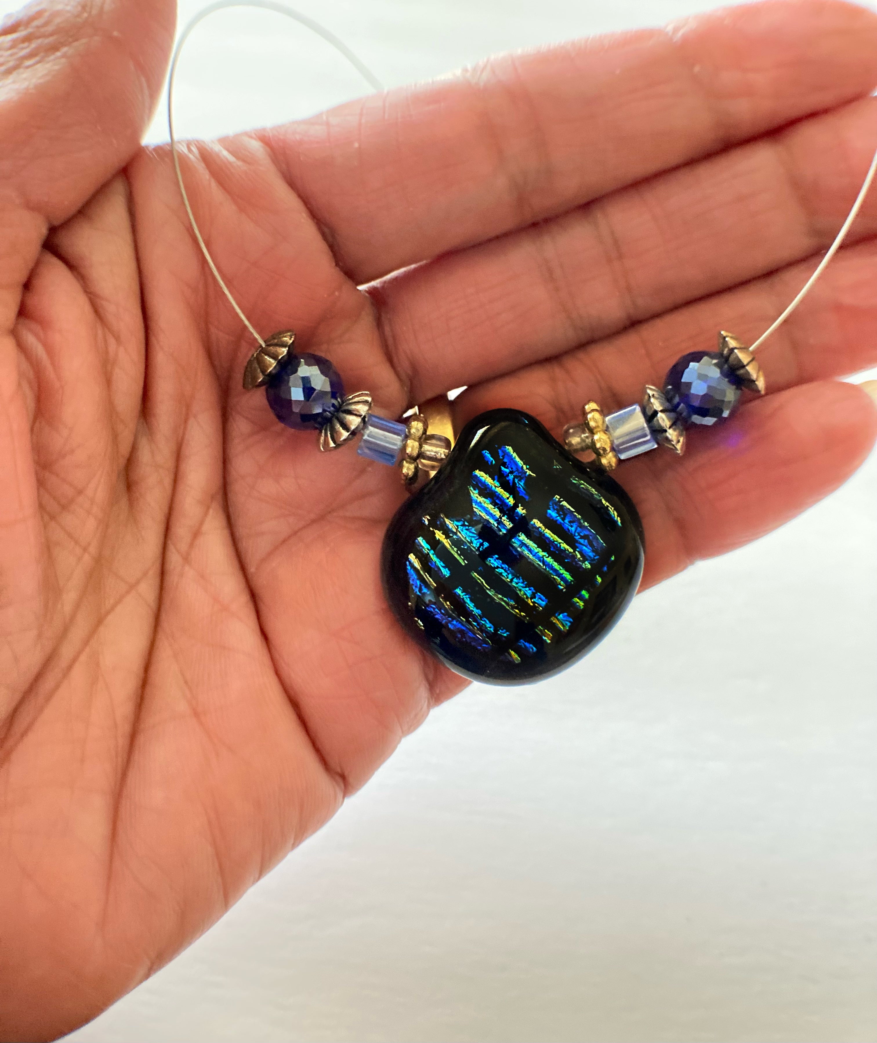 Hand holding beaded necklace with blue striped dichroic glass pendant against a white background, by 3D Glass Designs.