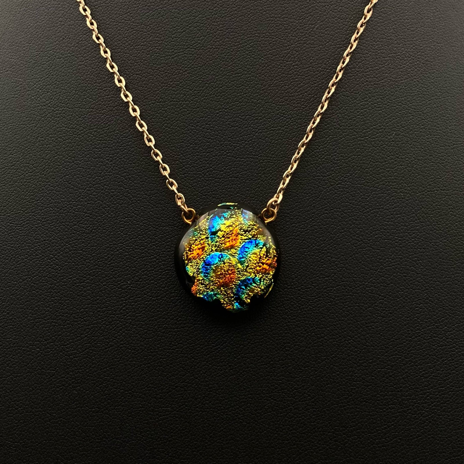 Colorful round fused glass pendant on a gold chain against a black leather background, by 3D Glass Designs.