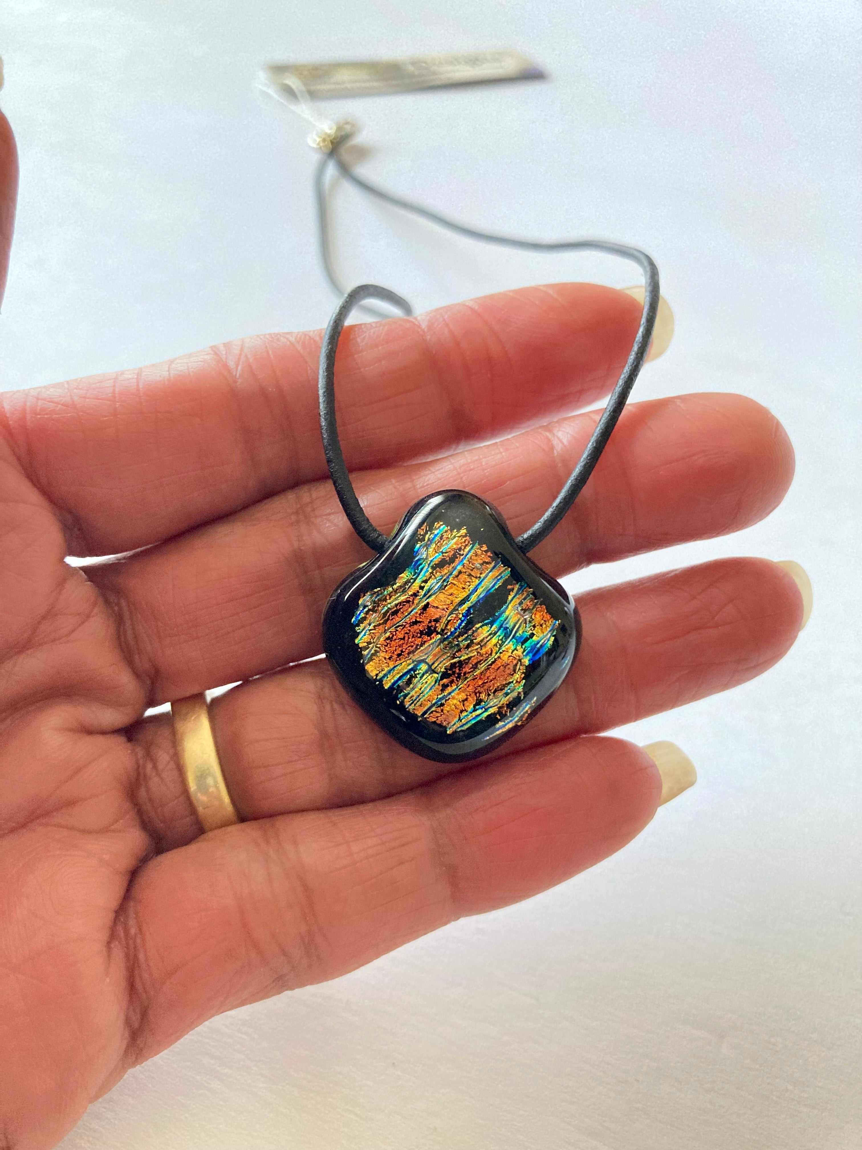 Models hand holding a colorful glass pendant on a black leather cord against a white background, by 3D Glass Designs.