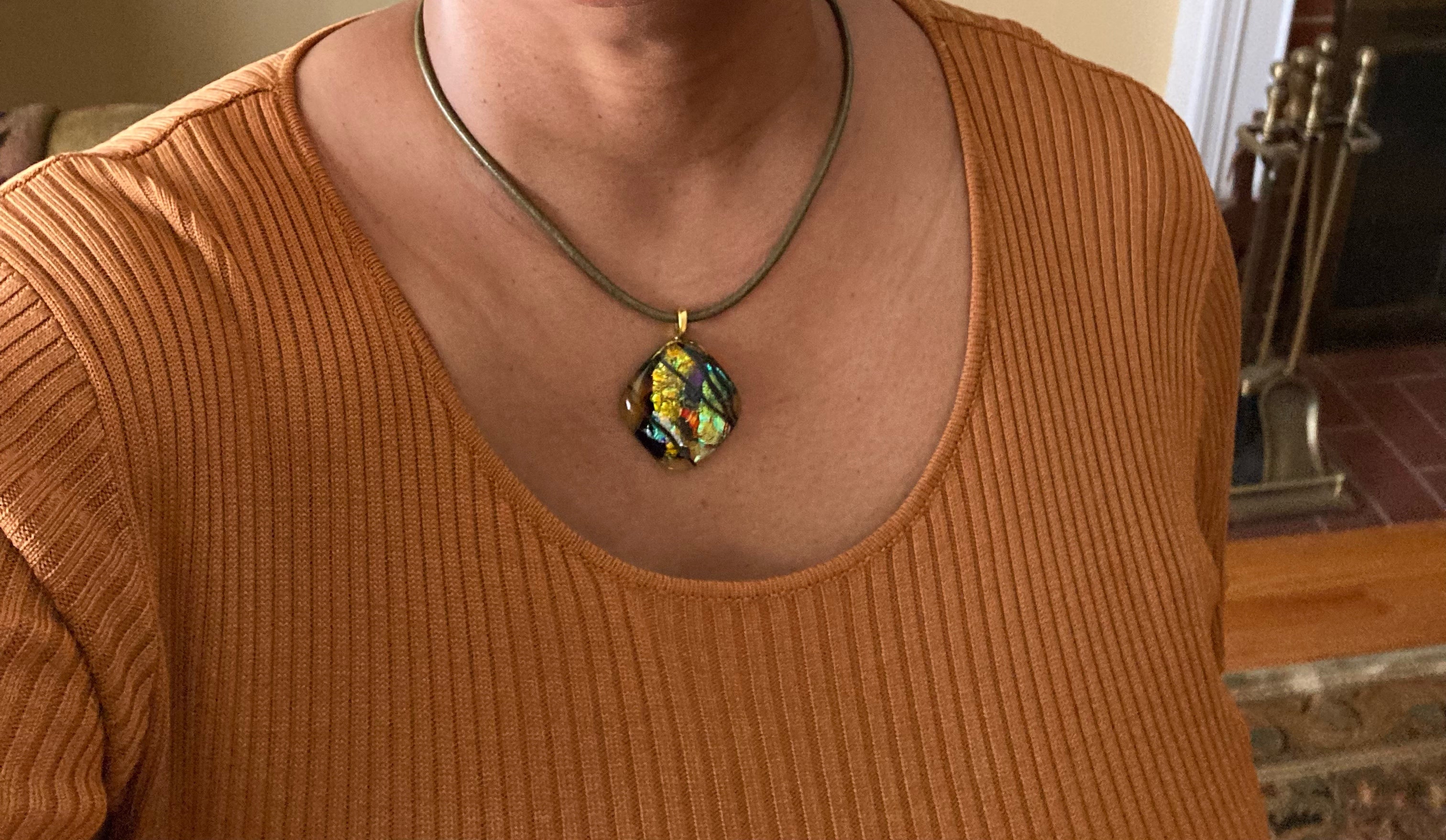 Model wearing fused glass pendant in transparent yellow with red, blue, green, purple and gold dichroic glass. Streaks of black add contrast to the design. Diamond shaped pendant hangs on an olive-green leather cord, by 3D Glass Designs.