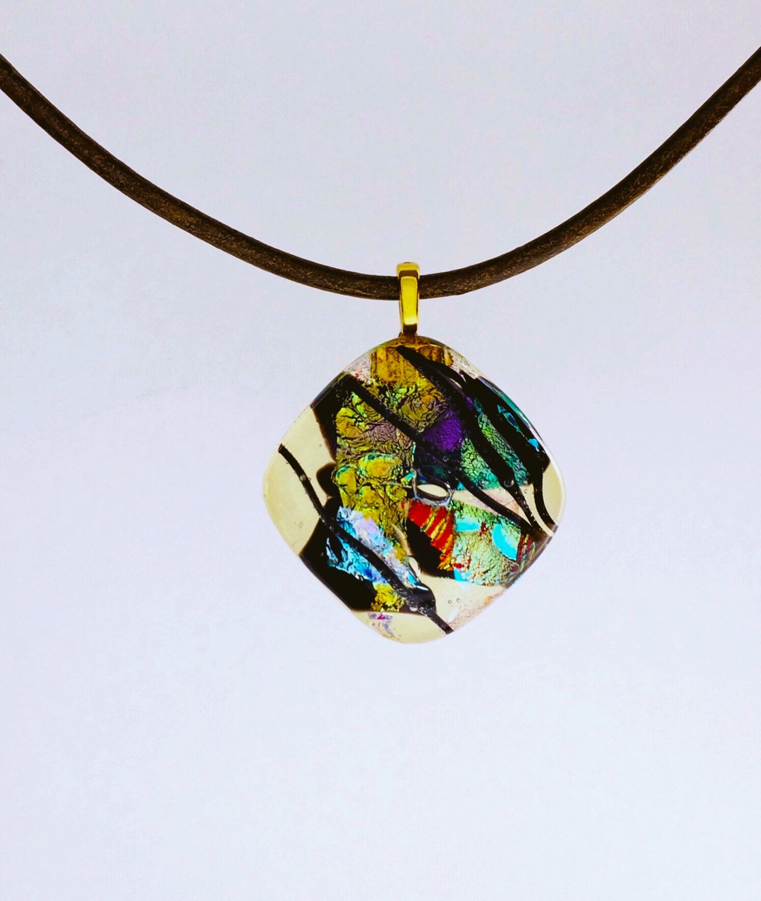 Fused glass pendant in transparent yellow with red, blue, green, purple and gold dichroic glass. Streaks of black add contrast to the design. Diamond shaped pendant hangs on an olive-green leather cord, by 3D Glass Designs.
