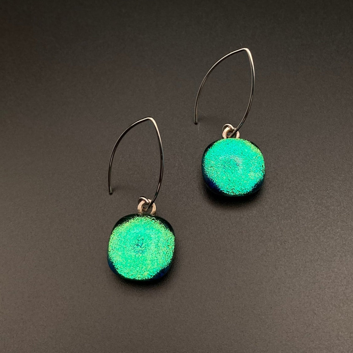 A pair or pierced earrings with teal colored glass dangles against a grey background, by 3D Glass Designs.