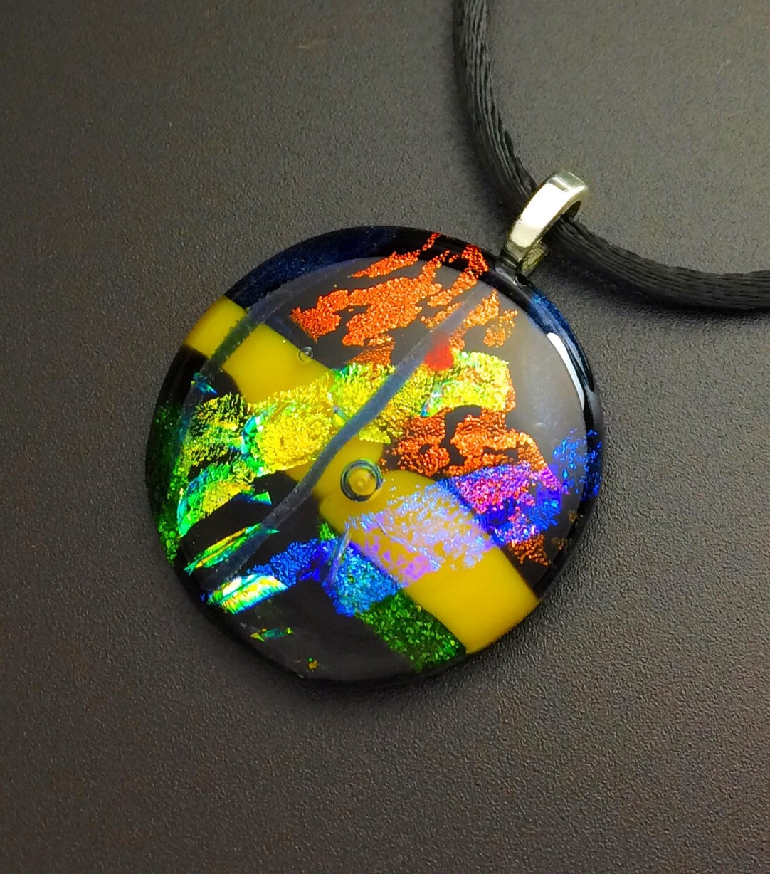 Round fused glass pendant necklace in green, yellow, blue, and rust hung on a 16-inch satin cord, by 3D Glass Designs.