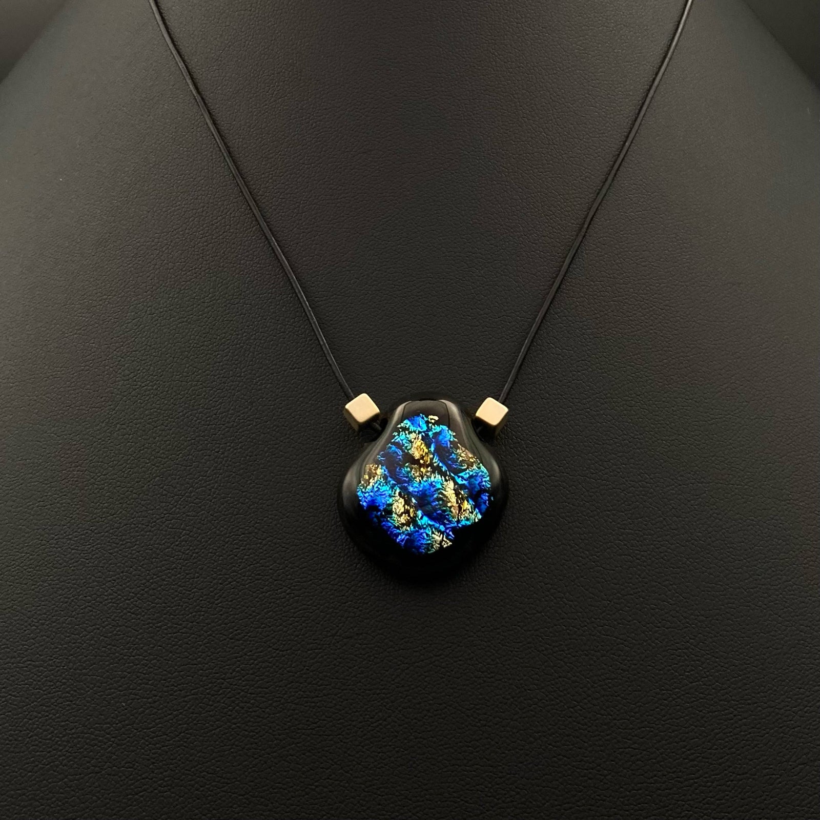 Small pear-shaped glass pendant with gold cube beads on a black leather cord against a black background, by 3D Glass Designs.