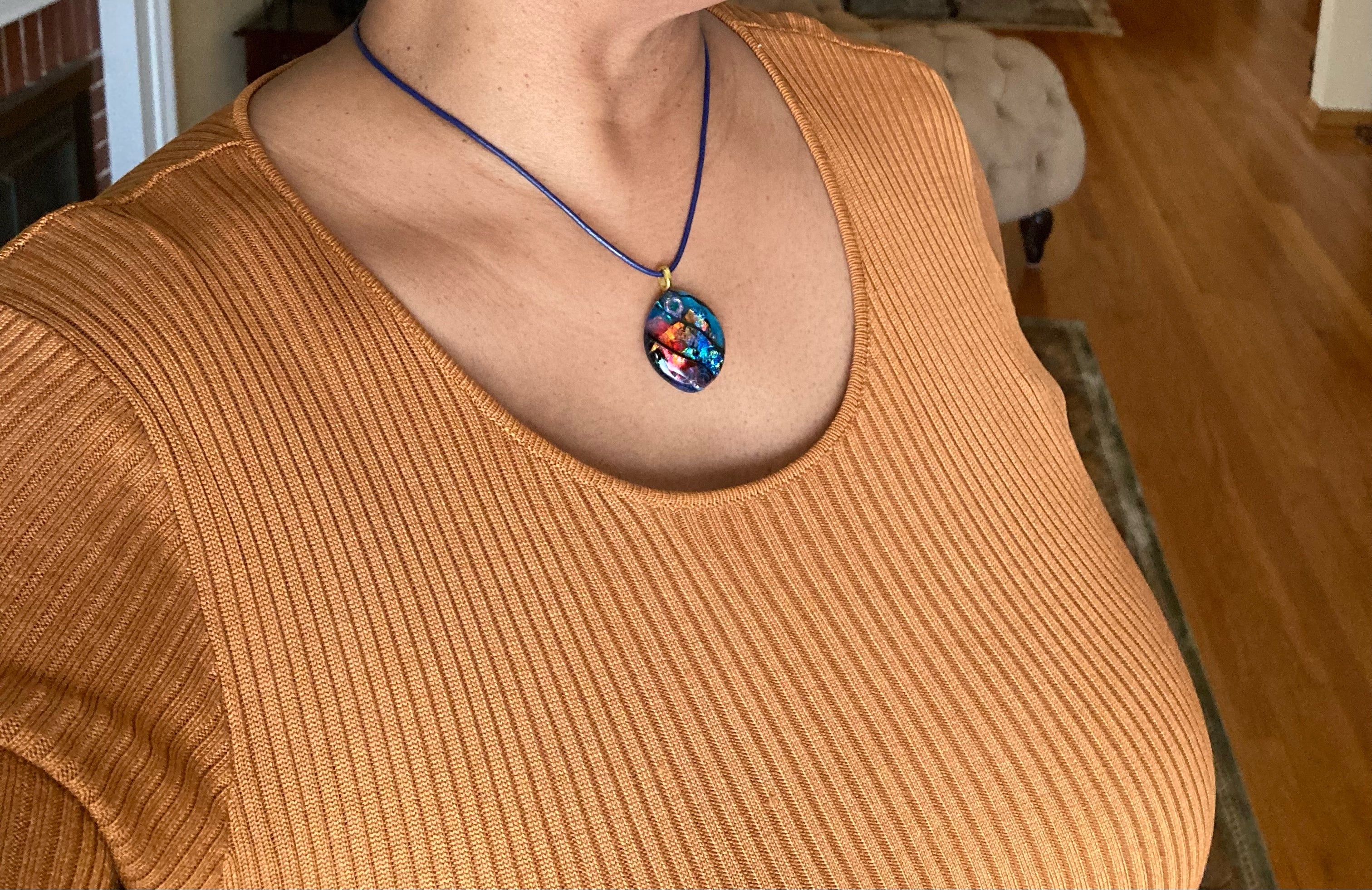 Model wearing fused glass pendant on a blue leather cord. Transparent blue glass with layers of red, rust, blue, green and purple dichroic glass and a copper squiggle design embedded in the glass. By 3D Glass Designs