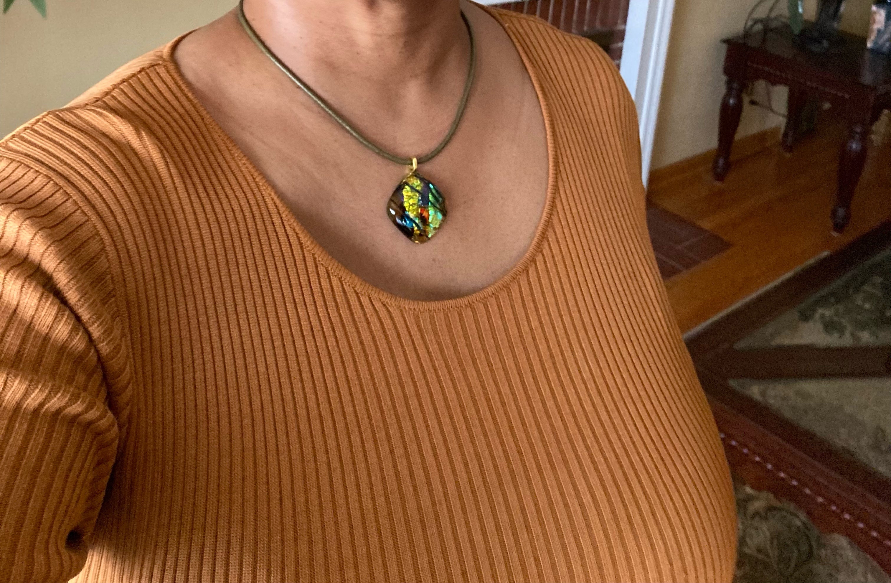 Model wearing fused glass pendant in transparent yellow with red, blue, green, purple and gold dichroic glass. Streaks of black add contrast to the design. Diamond shaped pendant hangs on an olive-green leather cord, by 3D Glass Designs.