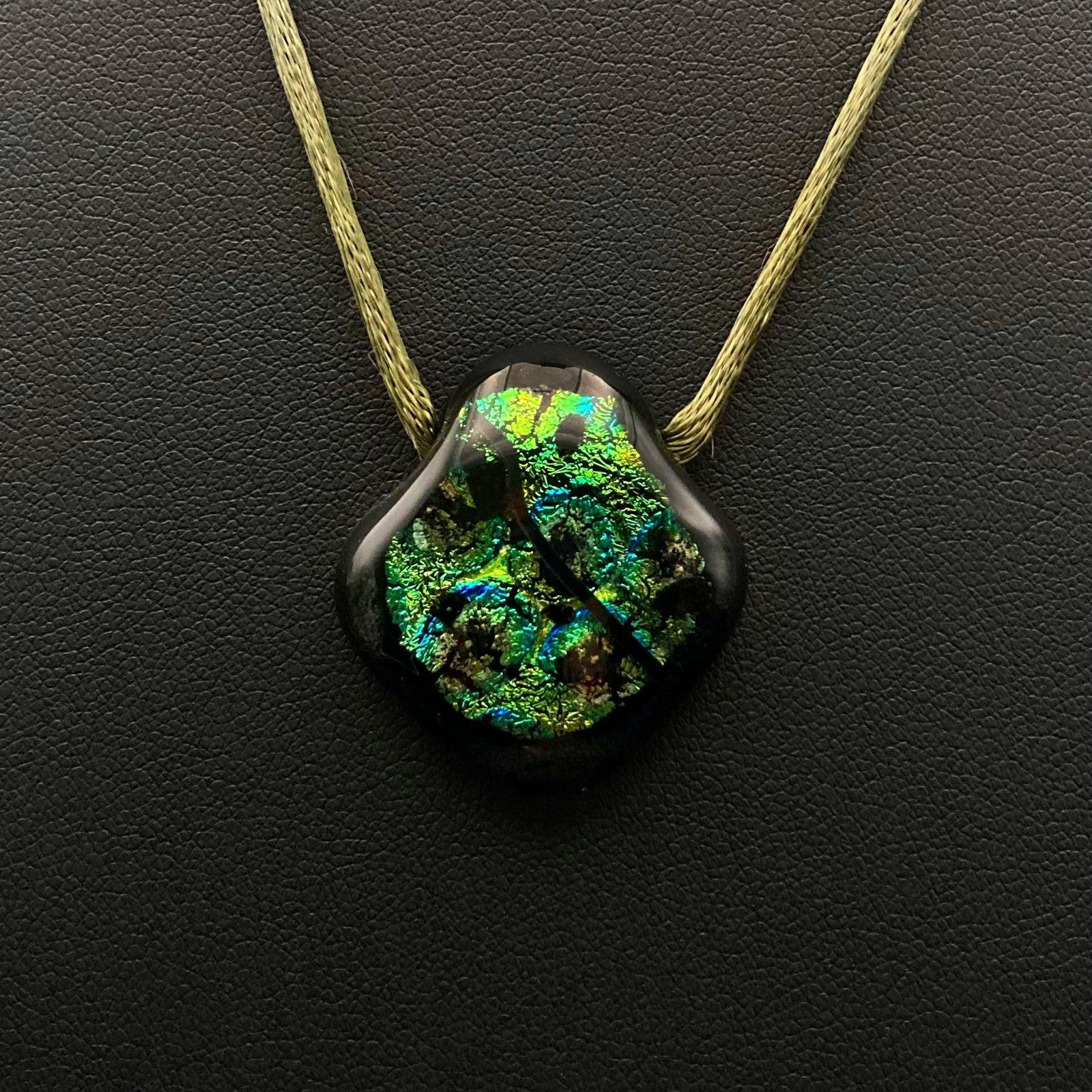 Green glass pendant on an olive-green satin cord against a black leather background, by 3D Glass Designs.