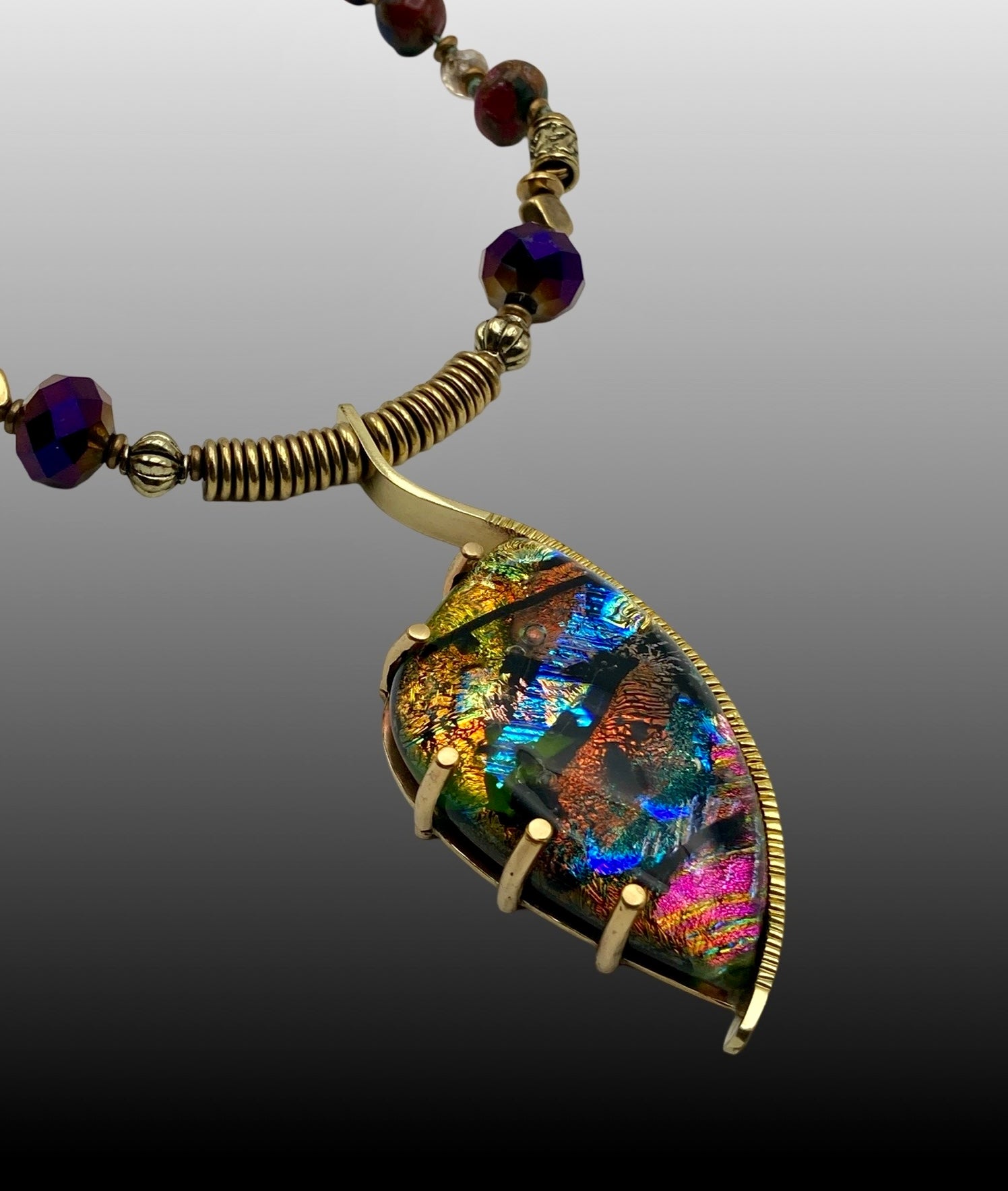 Dichroic glass set in a unique brass pendant, with prongs on left side and a textured bezel on the right side. Pendant is hung on a 29" long hand-knotted multicolor beaded necklace, by 3D Glass Designs.