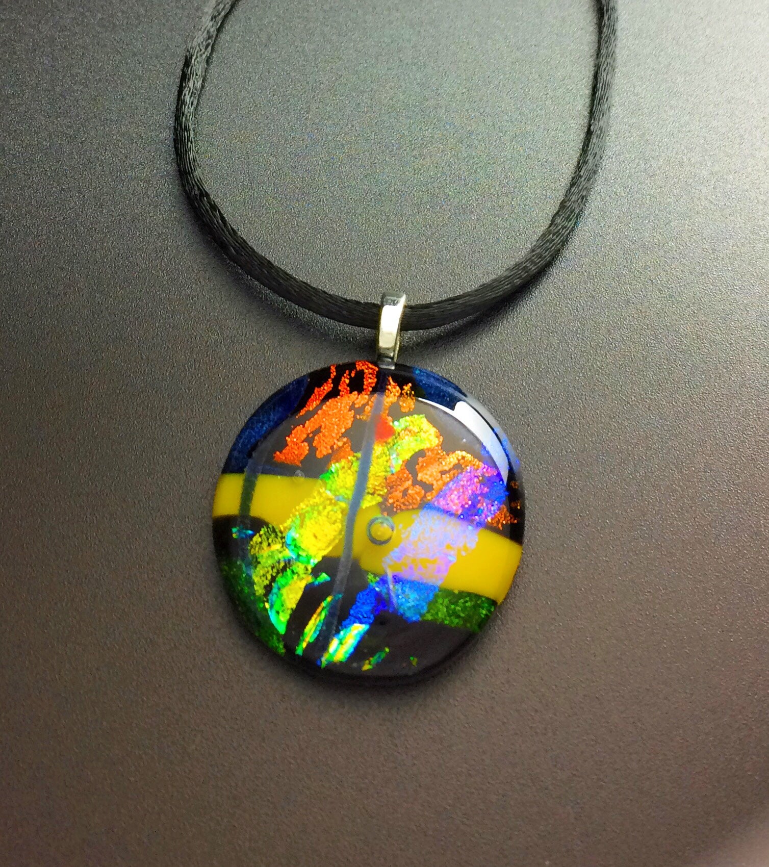 Round fused glass pendant necklace in green, yellow, blue, and rust hung on a 16-inch satin cord, by 3D Glass Designs.