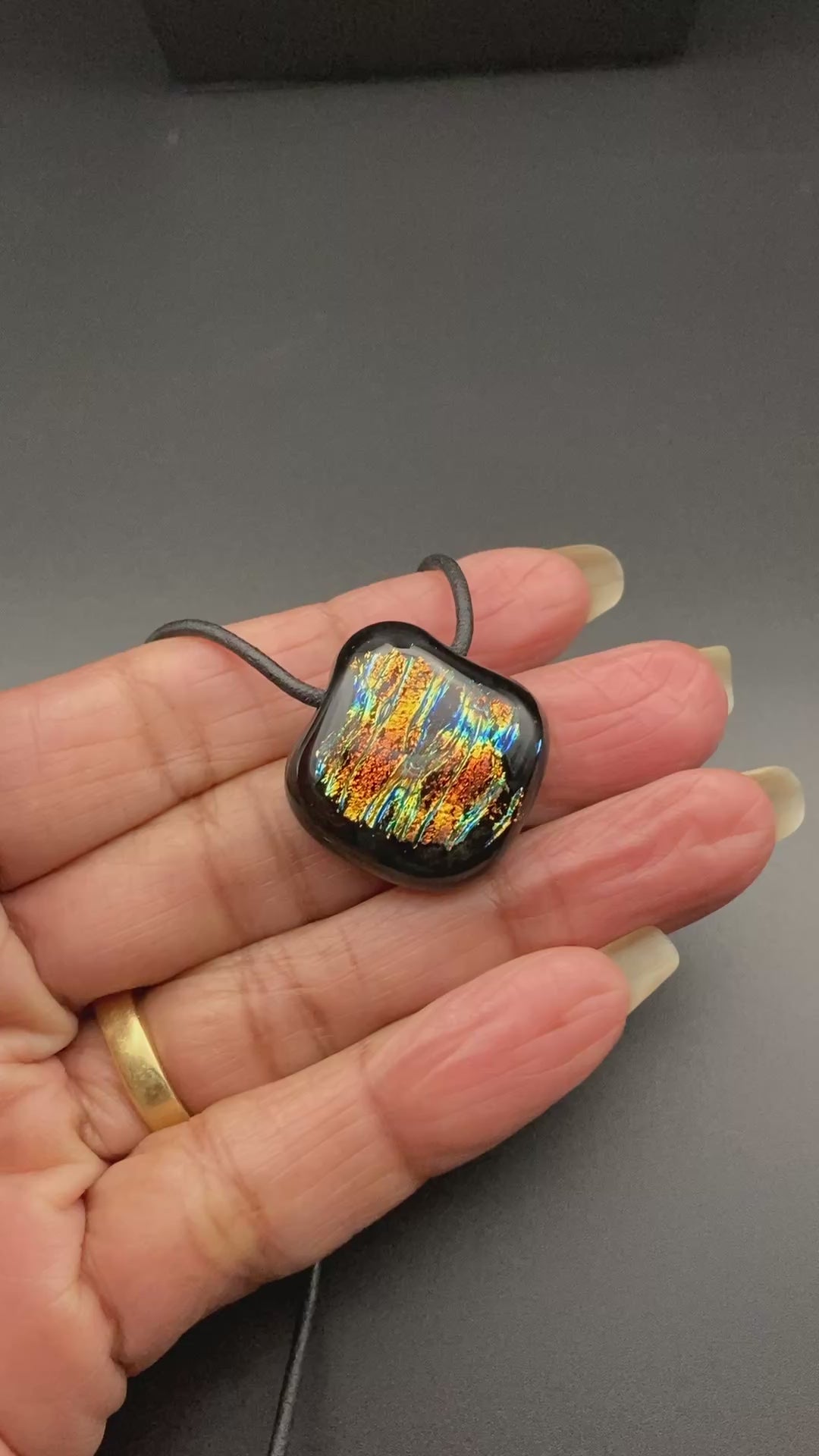 Hand holding a small multi-colored fused glass pendant on a black leather cord, by 3D lass Designs.