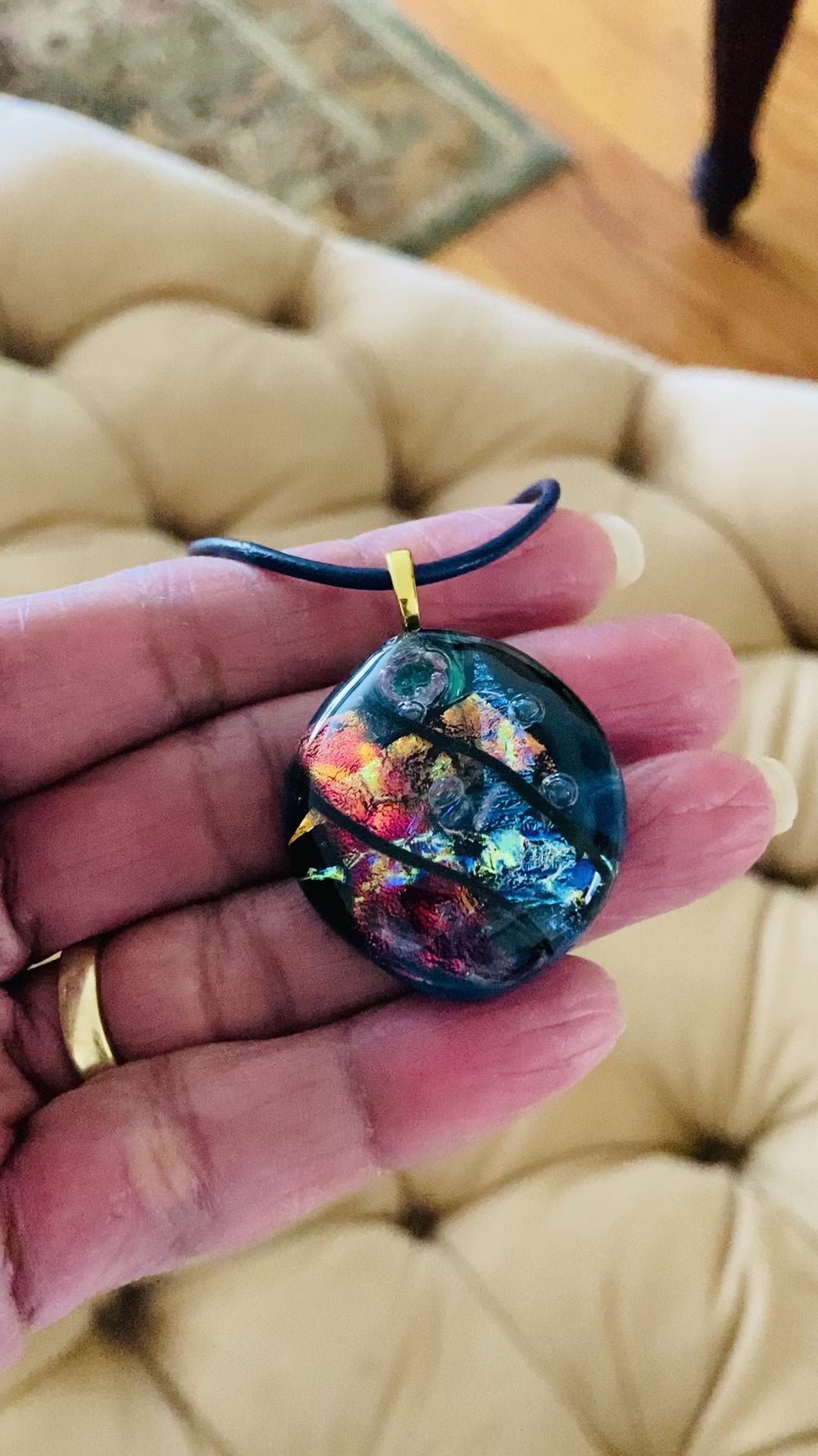 Video of hand holding fused glass pendant on a blue leather cord. Transparent blue glass with layers of red, rust, blue, green and purple dichroic glass and a copper squiggle design embedded in the glass. By 3D Glass Designs