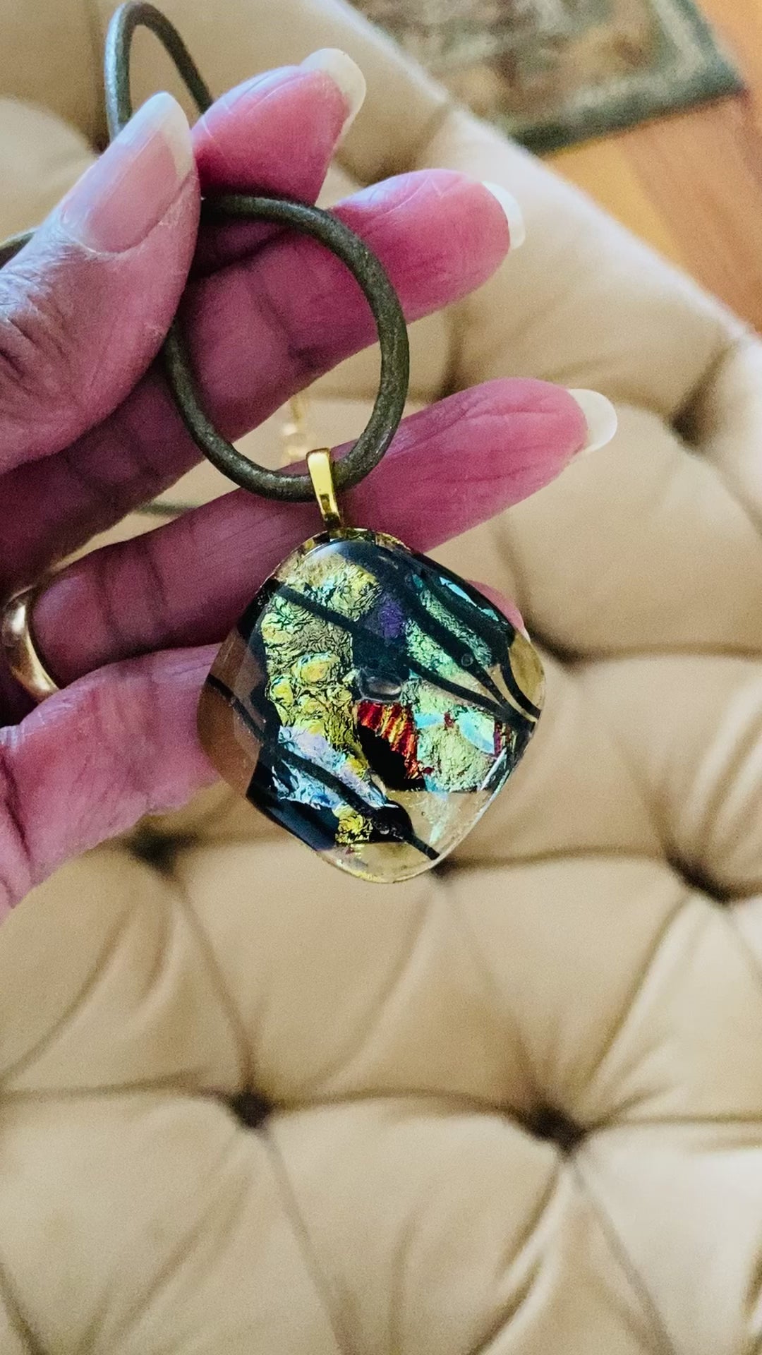 Video of hand holding fused glass pendant in transparent yellow with red, blue, green, purple and gold dichroic glass. Streaks of black add contrast to the design. Diamond shaped pendant hangs on an olive-green leather cord, by 3D Glass Designs.