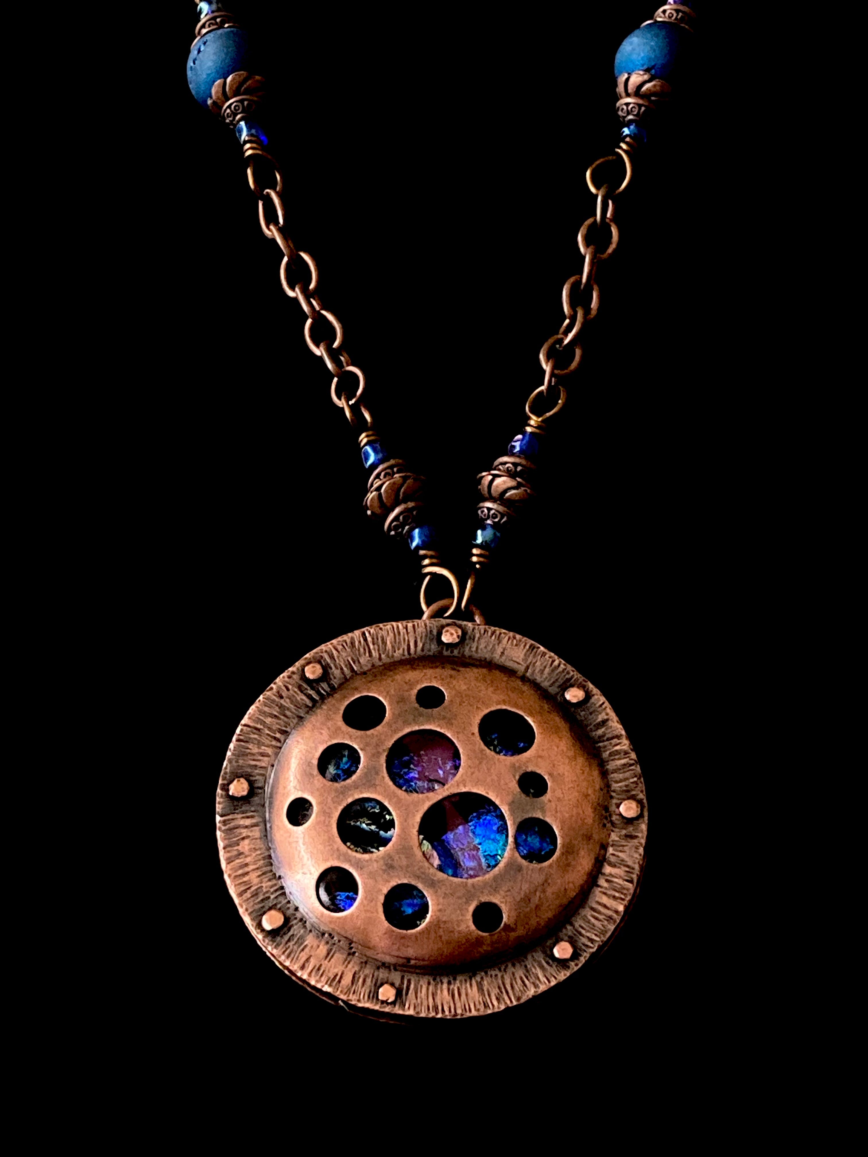 Round copper pendant with various sized holes revealing pink and blue fused glass beneath, sandwiched between two layers of copper. This unique artisan pendant is hung from a beaded copper chain, by 3D Glass Designs.
