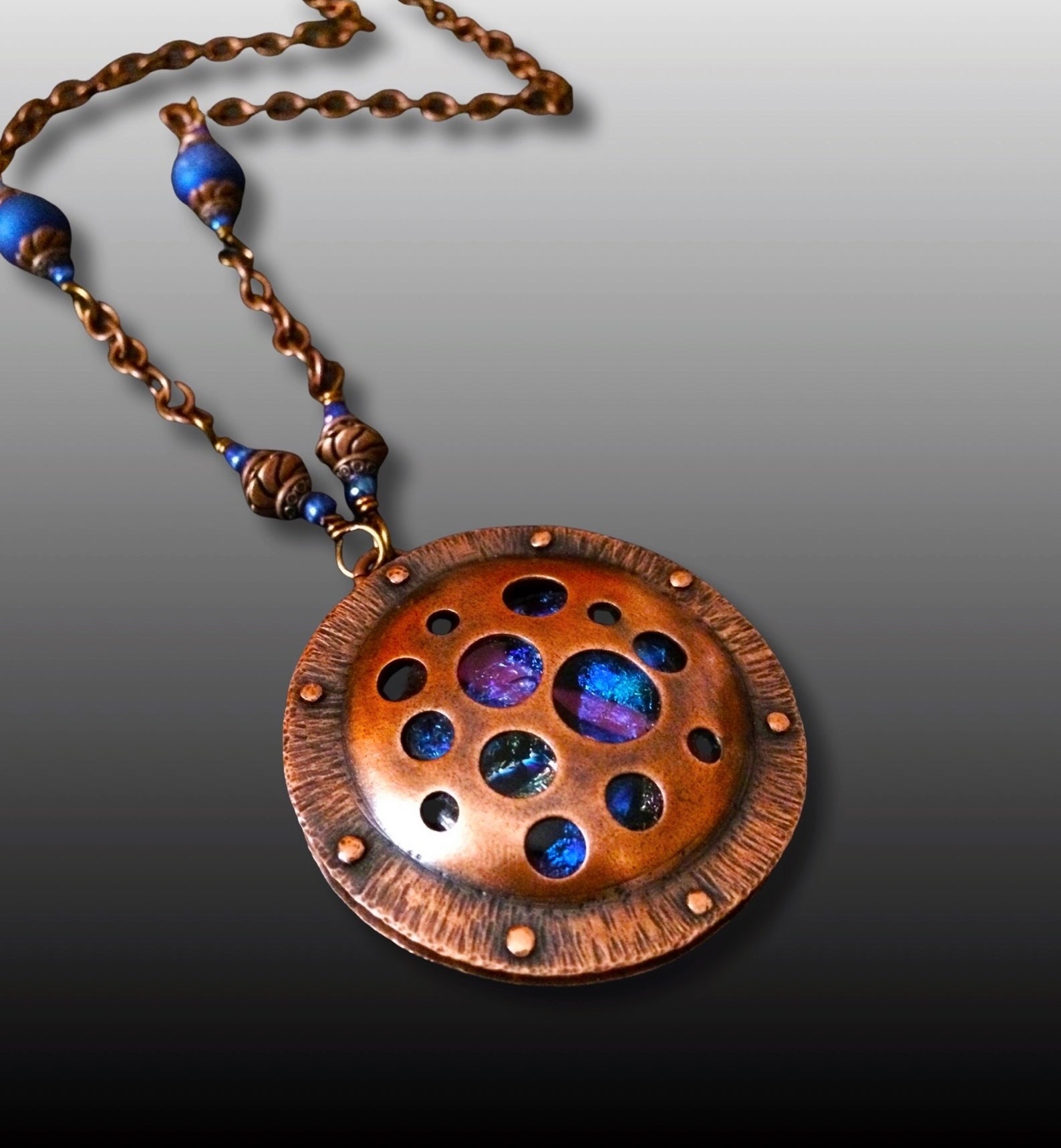 Round copper pendant with various sized holes revealing pink and blue fused glass beneath, sandwiched between two layers of copper. This unique artisan pendant is hung from a beaded copper chain, by 3D Glass Designs.