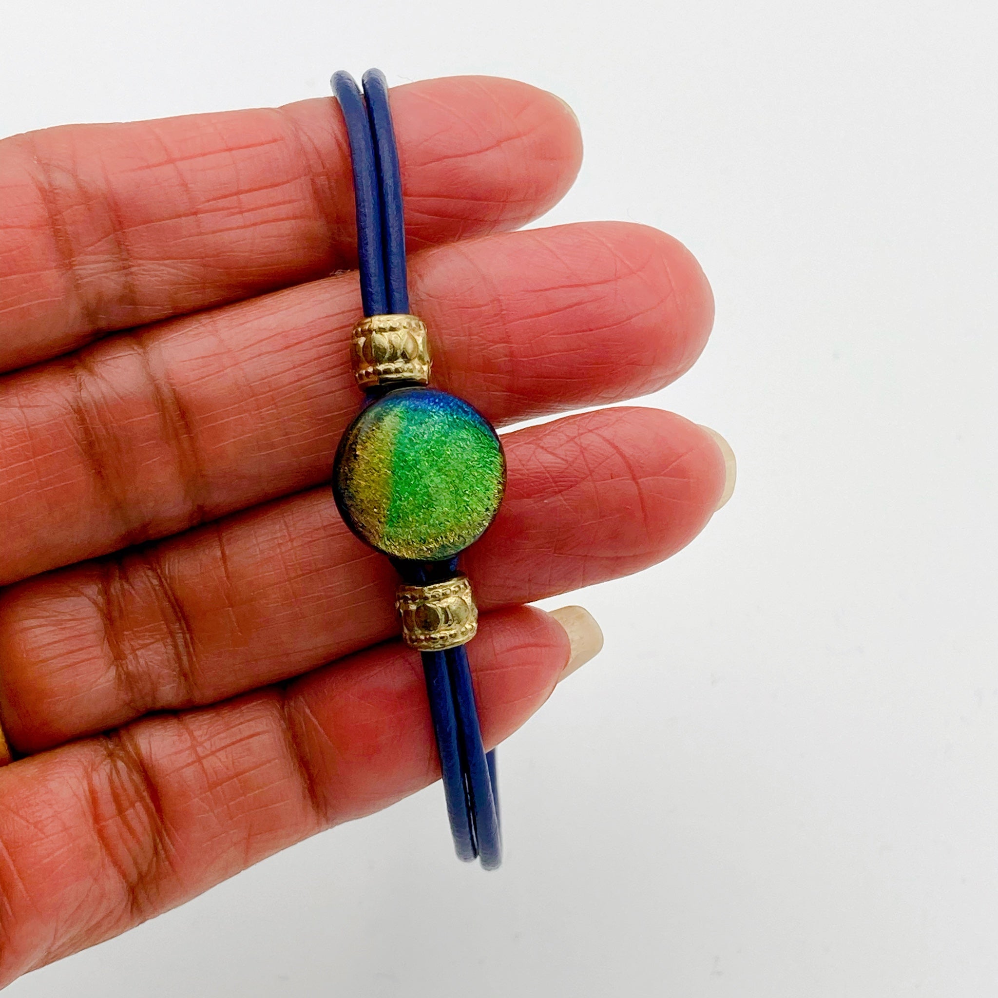 Hand holding handmade blue leather bracelet with green dichroic glass, by 3D Glass Designs.