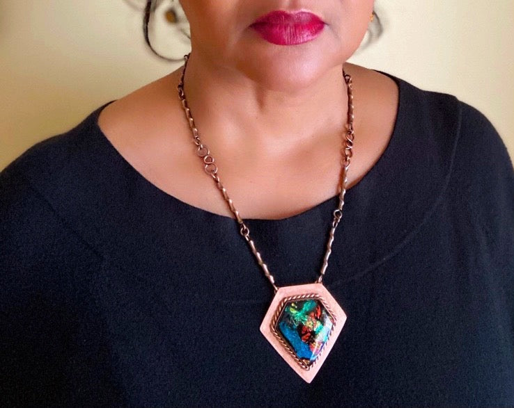 Model wearing one-of-a-kind fused glass and copper pendant necklace by 3D Glass Designs.  Dichroic glass in blue, green and rust hues set in a handcrafted diamond-shaped shield copper pendant with an elegant rope detail surrounding the bezel.  The handmade copper necklace consists of twisted copper links that resembles bamboo and a handmade clasp.