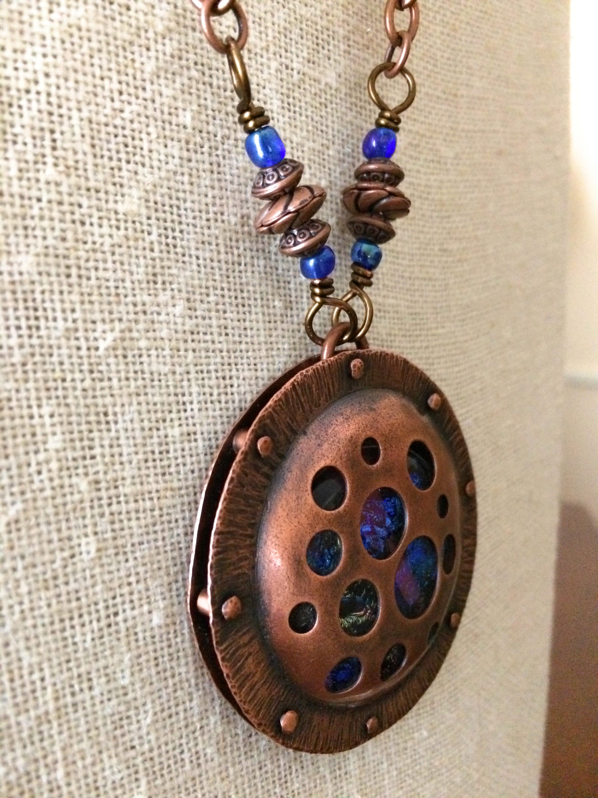 Round copper pendant with various sized holes revealing pink and blue fused glass beneath, sandwiched between two layers of copper. This unique artisan pendant is hung from a beaded copper chain, by 3D Glass Designs.
