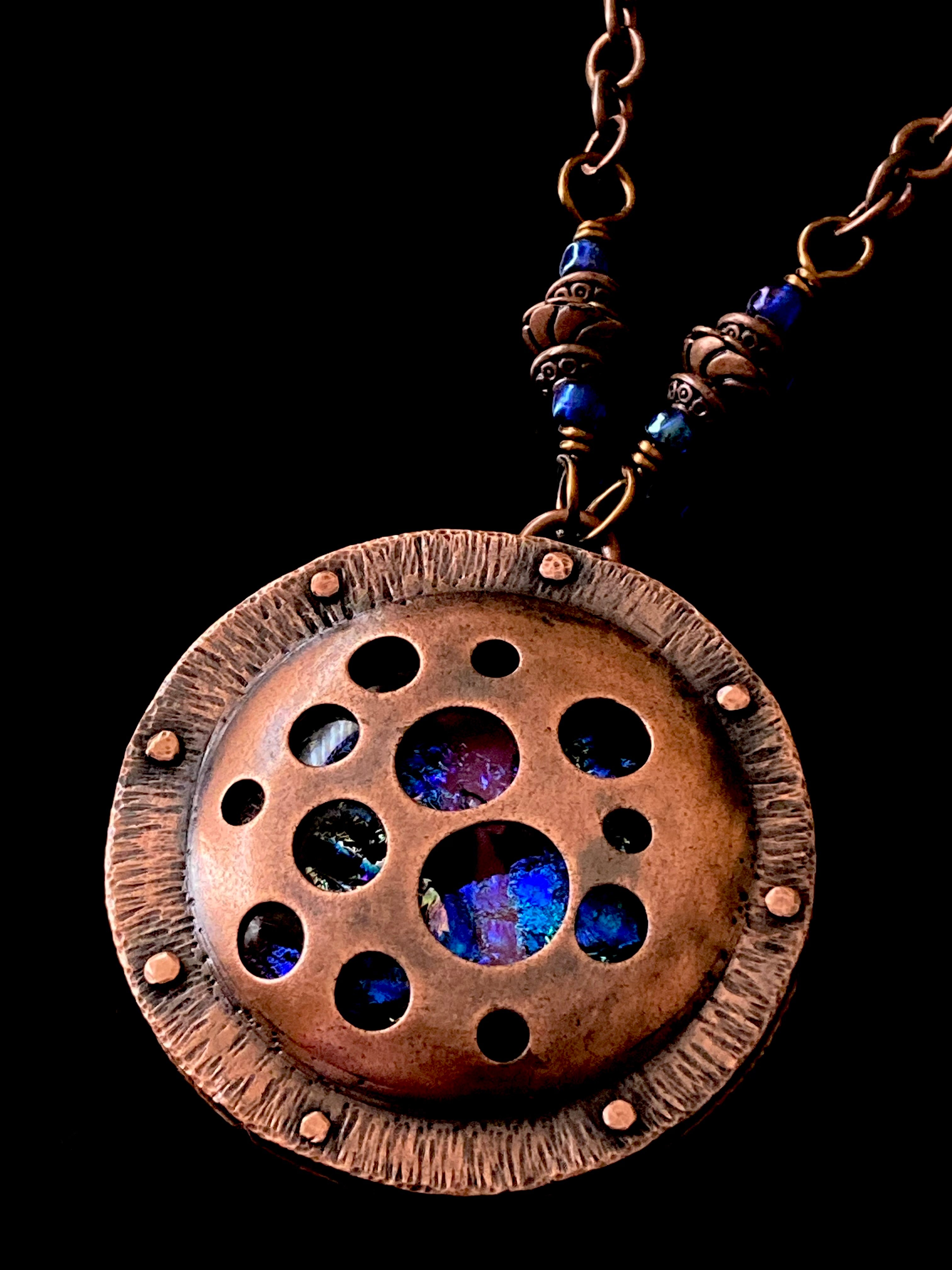 Round copper pendant with various sized holes revealing pink and blue fused glass beneath, sandwiched between two layers of copper. This unique artisan pendant is hung from a beaded copper chain, by 3D Glass Designs.