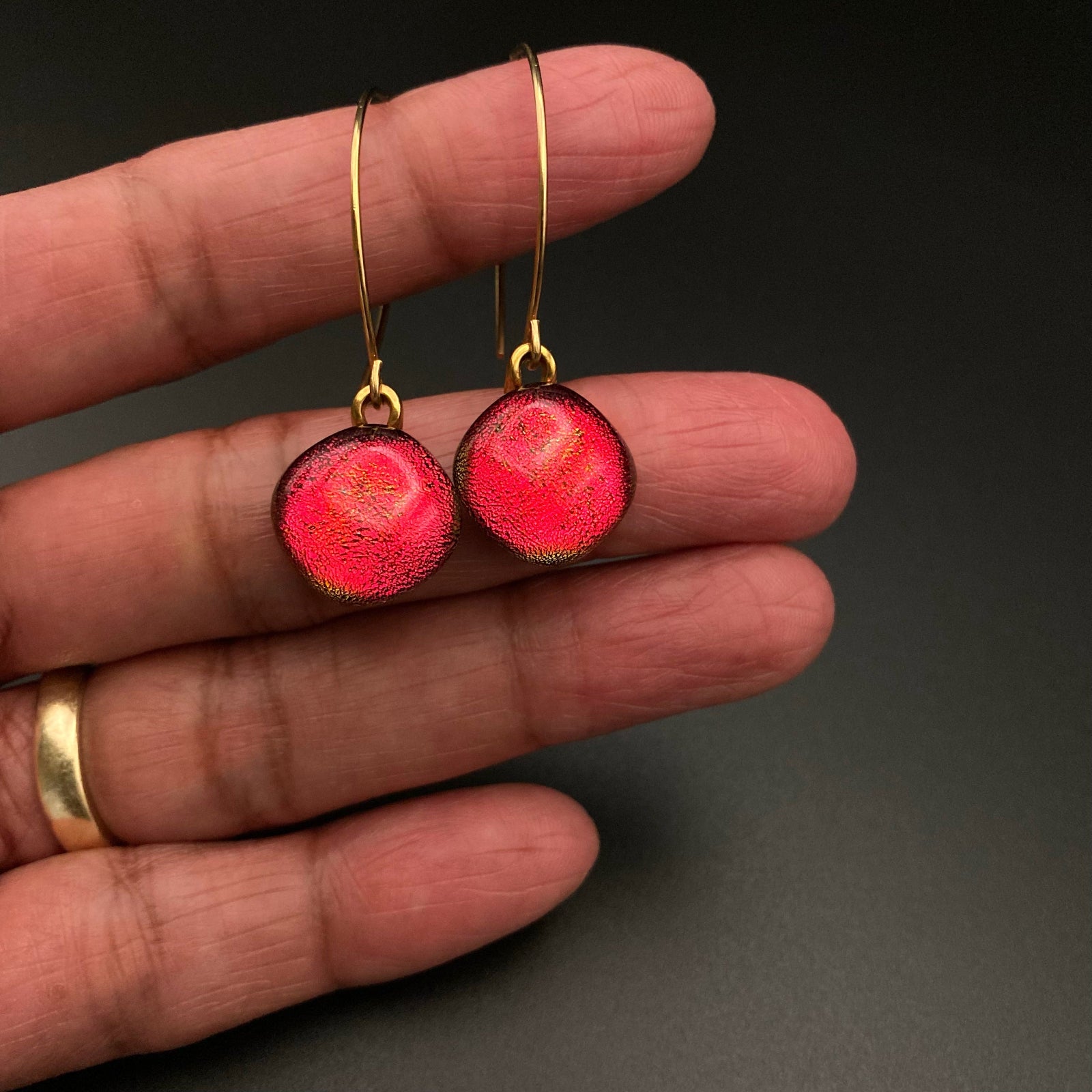 Hand holding a pair of red glass earrings on long gold ear wires, by 3D Glass Designs.