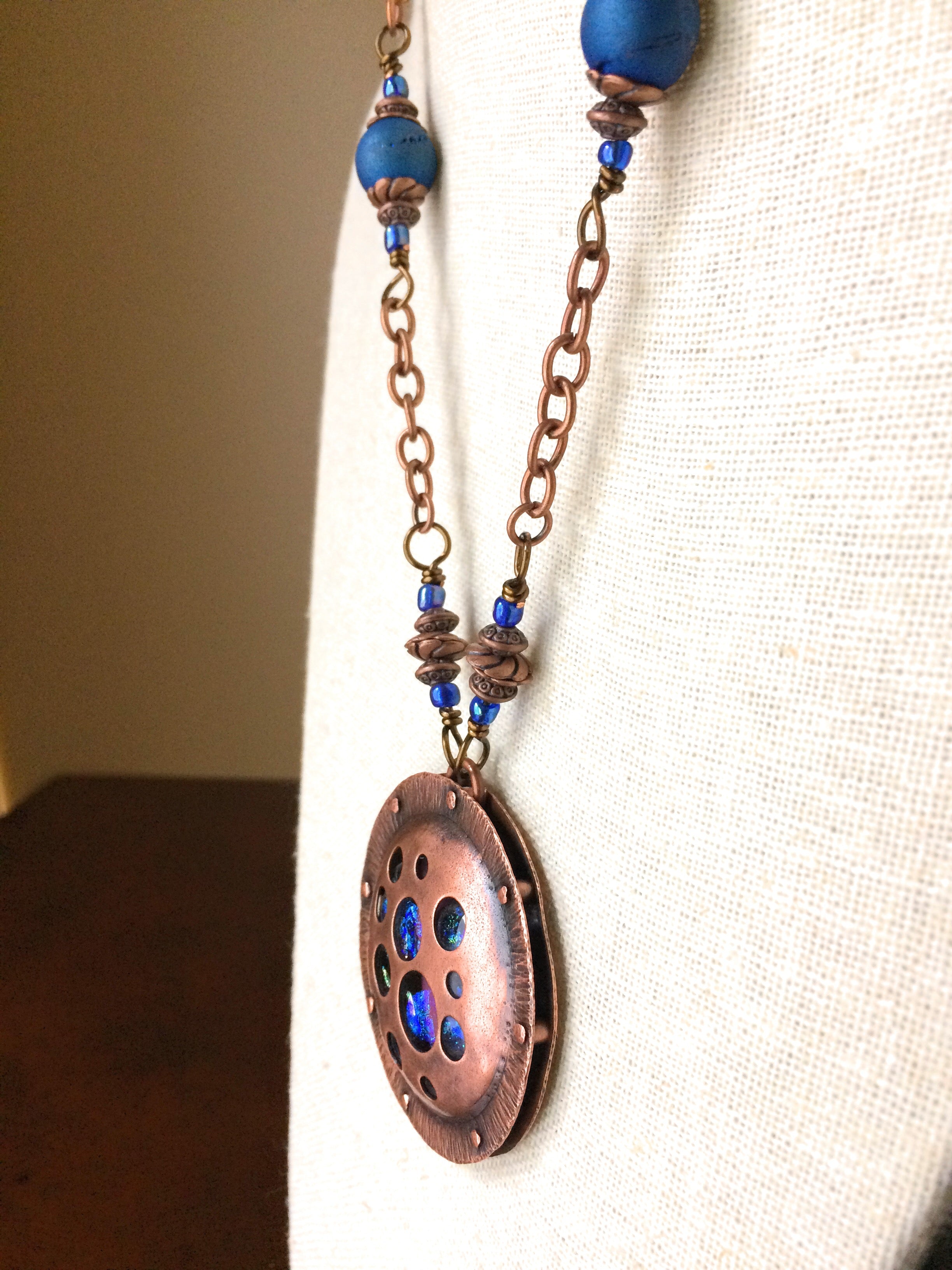 Round copper pendant with various sized holes revealing pink and blue fused glass beneath, sandwiched between two layers of copper. This unique artisan pendant is hung from a beaded copper chain, by 3D Glass Designs.
