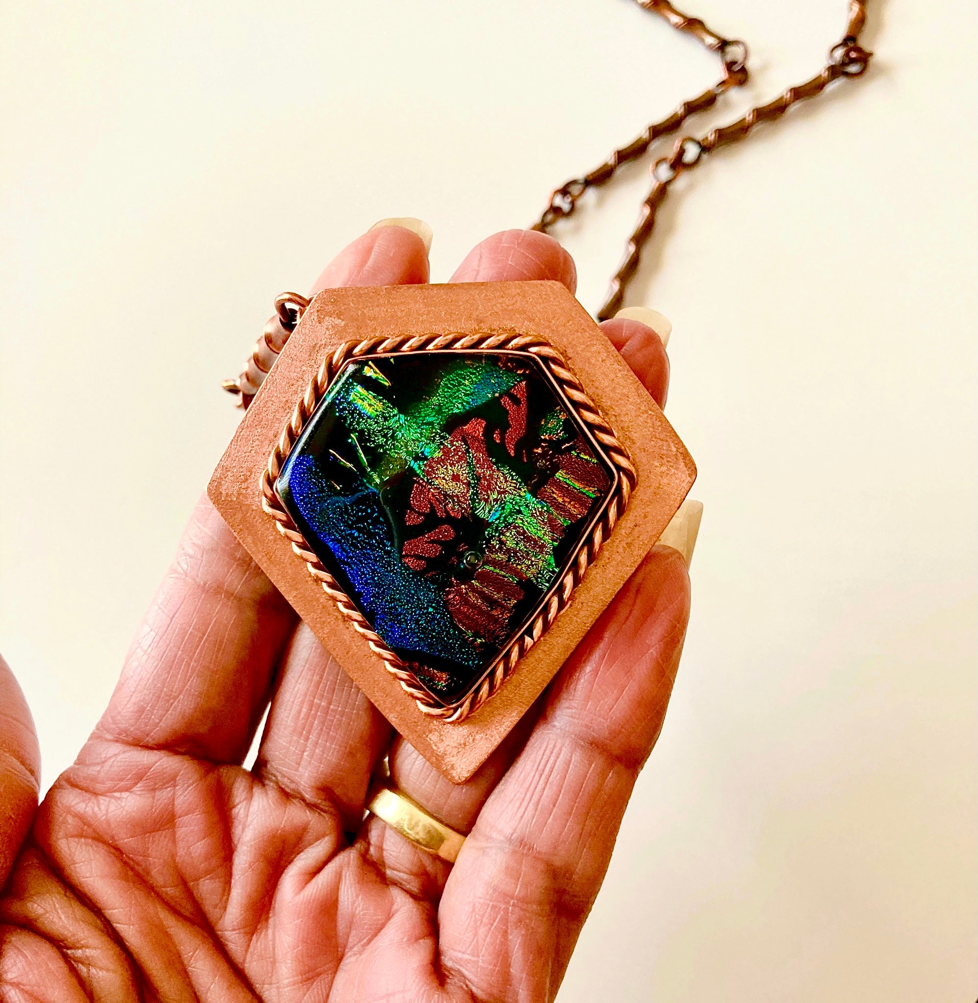 Model's hand showing the details of fused glass and copper pendant necklace by 3D Glass Designs.  Dichroic glass in blue, green and rust hues set in a handcrafted diamond-shaped shield copper pendant with an elegant rope detail surrounding the bezel.  The handmade copper necklace consists of twisted copper links that resembles bamboo and a handmade clasp.
