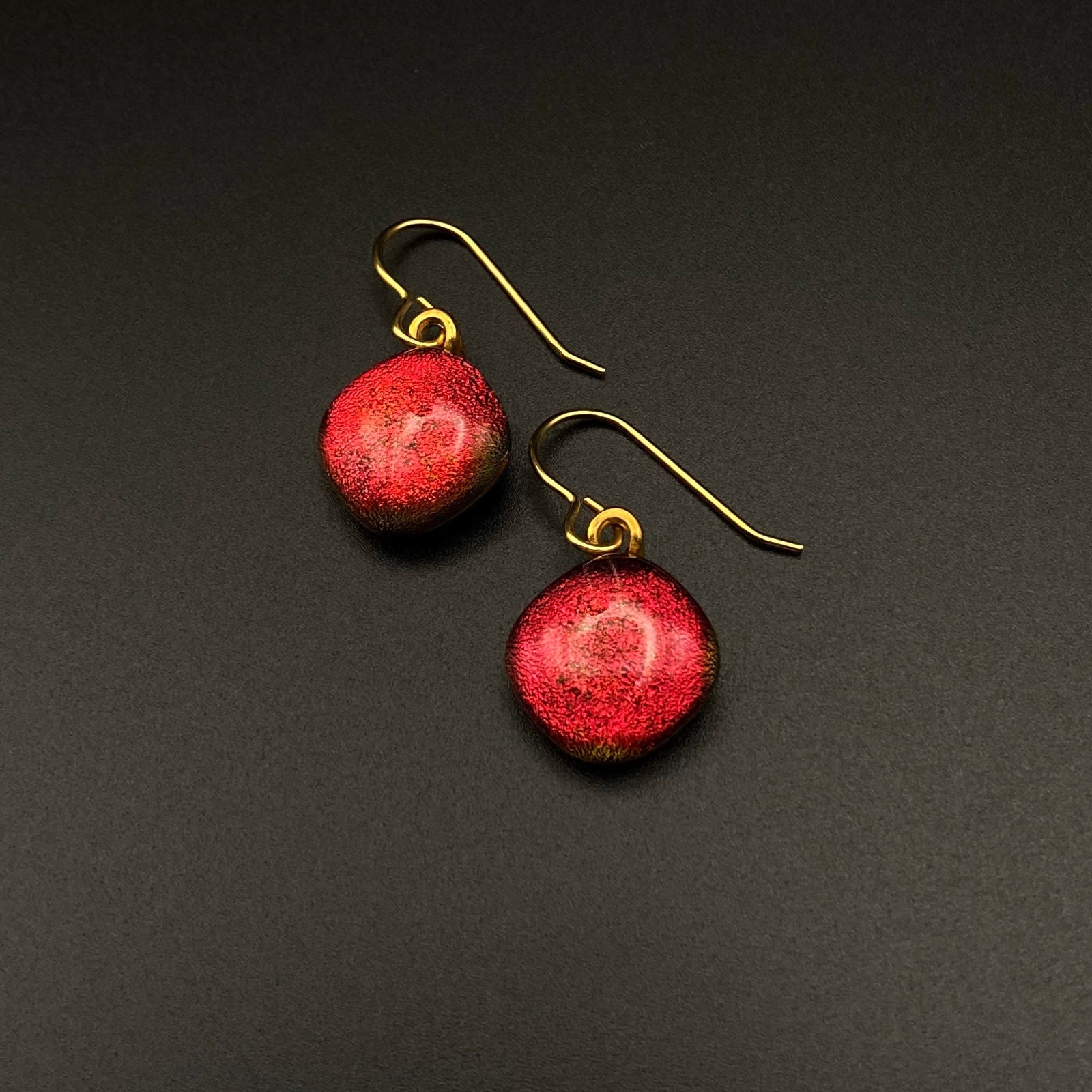 A pair of red glass earrings on gold ear wires against a grey background, by 3D Glass Designs.