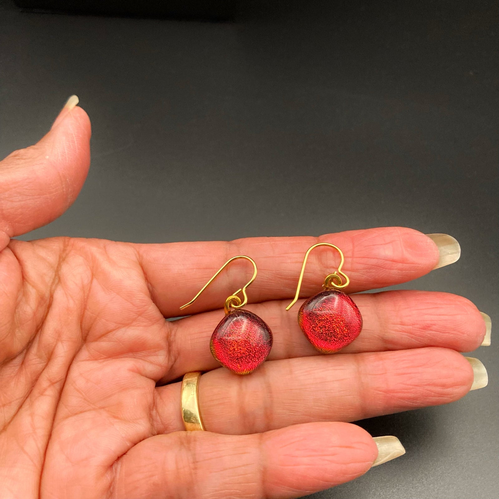 A hand holding a pair of red earrings on short gold ear wires, by 3D Glass Designs.