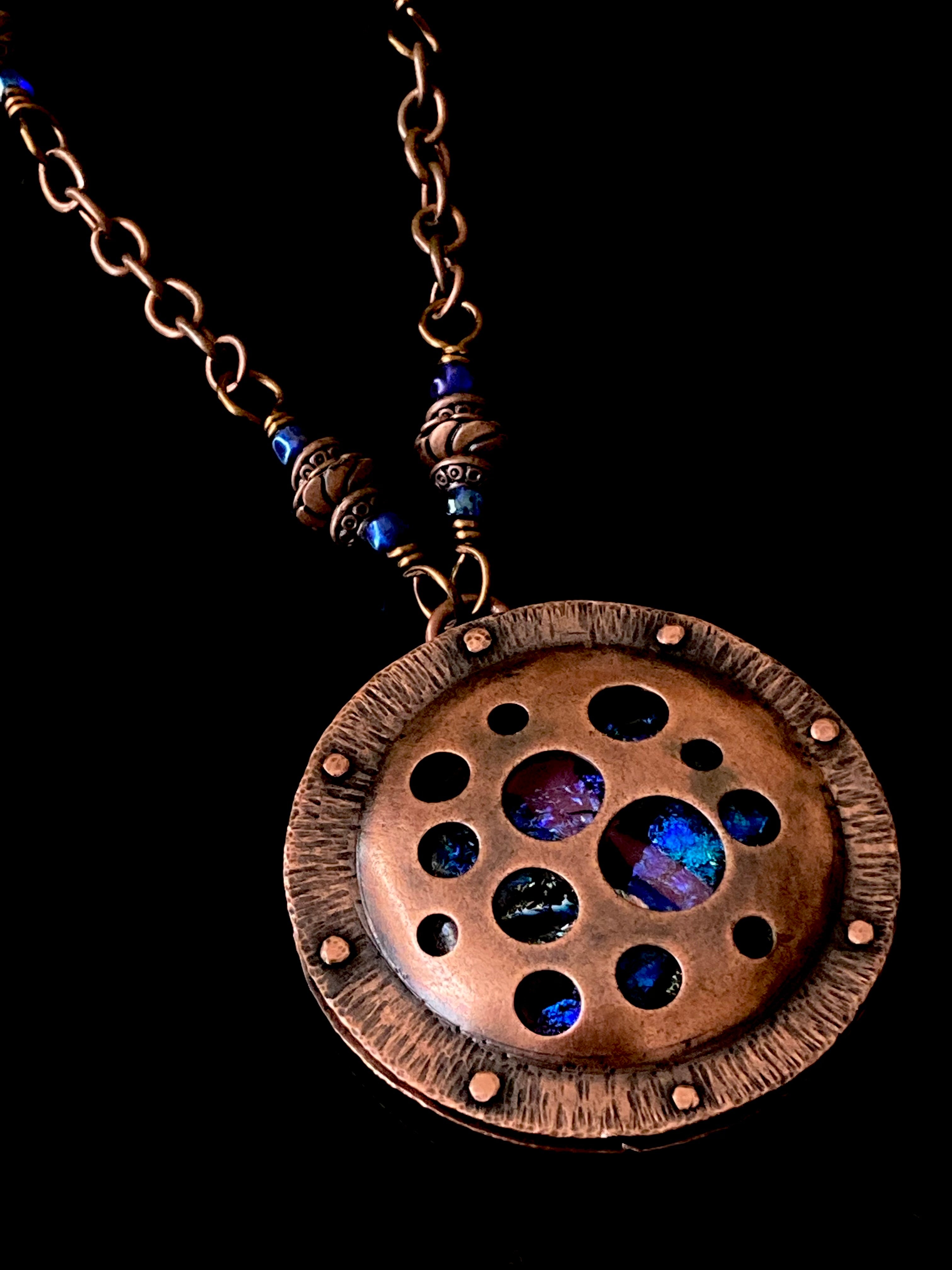 Round copper pendant with various sized holes revealing pink and blue fused glass beneath, sandwiched between two layers of copper. This unique artisan pendant is hung from a beaded copper chain, by 3D Glass Designs.