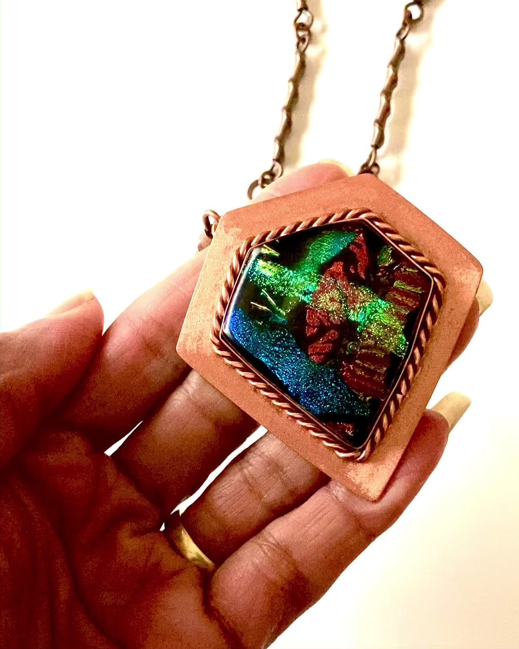 Video of model's hand showing the details of fused glass and copper pendant necklace by 3D Glass Designs.  Dichroic glass in blue, green and rust hues set in a handcrafted diamond-shaped shield copper pendant with an elegant rope detail surrounding the bezel.  The handmade copper necklace consists of twisted copper links that resembles bamboo and a handmade clasp.