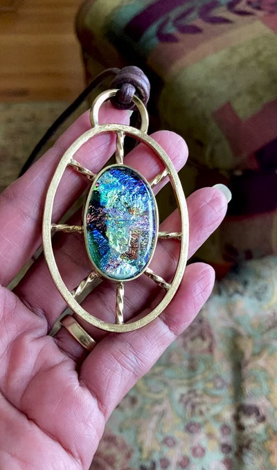 Video of hand holding Dichroic glass set in a unique brass pendant, with prongs on left side and a textured bezel on the right side. Pendant is hung on a 29" long hand-knotted multicolor beaded necklace, by 3D Glass Designs.