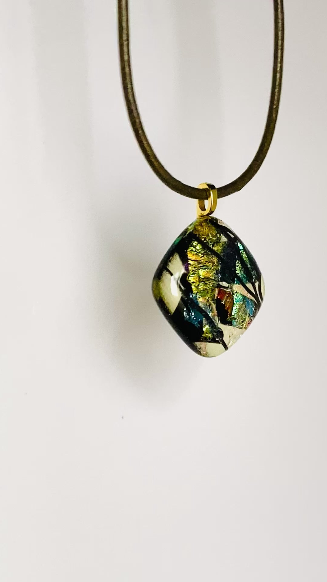 Video fused glass pendant in transparent yellow with red, blue, green, purple and gold dichroic glass. Streaks of black add contrast to the design. Diamond shaped pendant hangs on an olive-green leather cord, by 3D Glass Designs. 