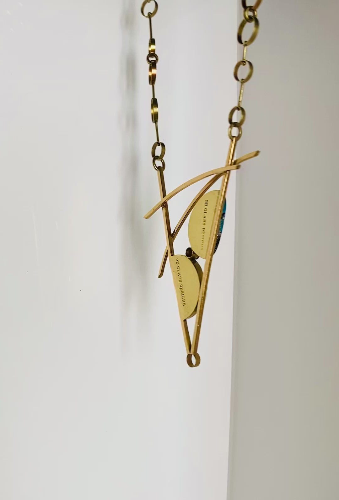 Video of geometric inspired brass and teal fused glass pendant necklace.  Large statement pendant with two glass semi-circles with a CZ in between, set in a V frame with two slightly cured arch's above.  Chain has various sized brass circles linked together, by 3D Glass Designs.