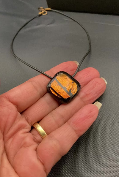 Hand holding orange and black fused glass pendant against a dark cloth background, by 3D Glass Designs.